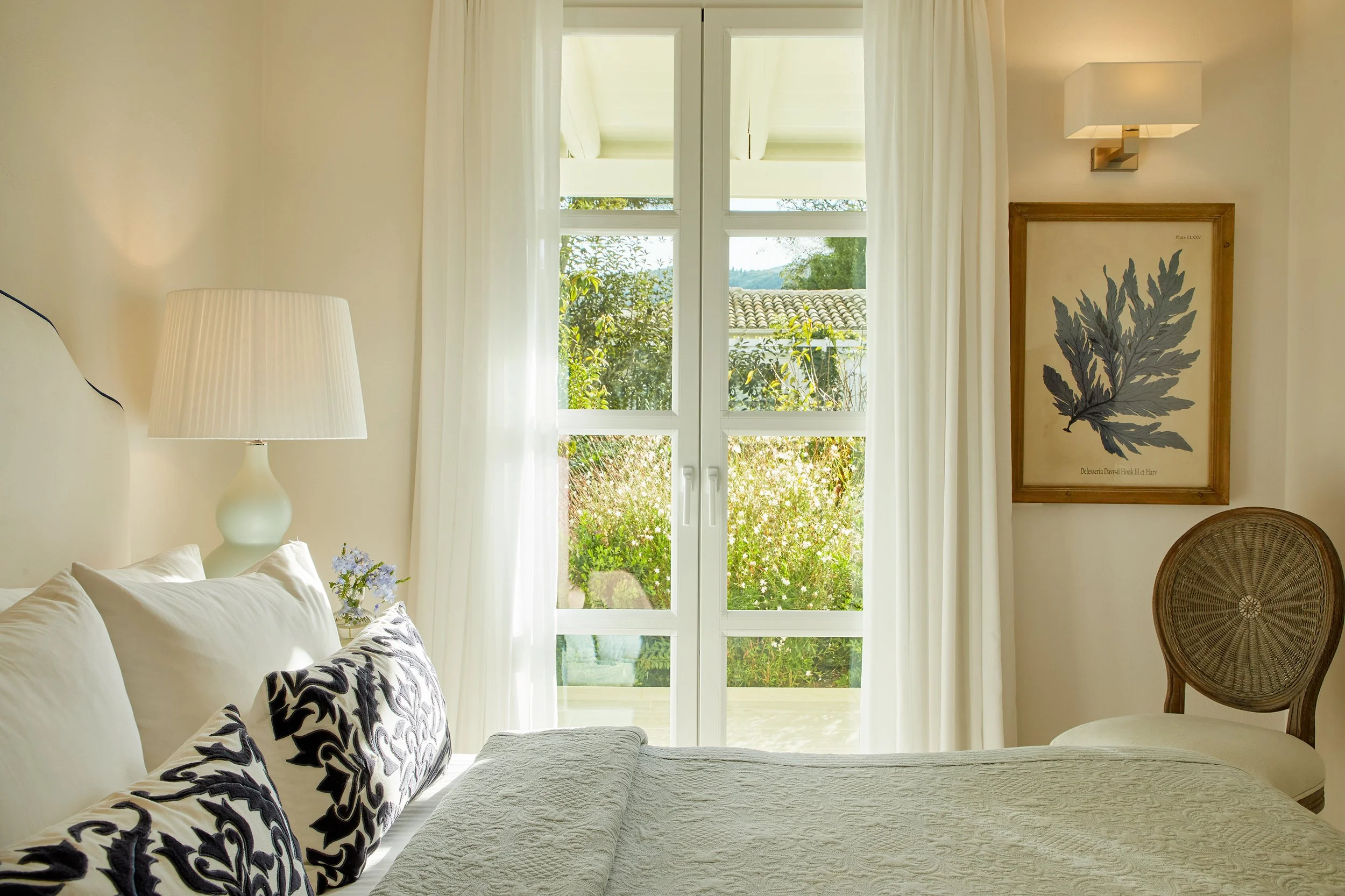 Bright bedroom with white curtains, a large window showing greenery outside, a bed with white and patterned pillows, a white bedside lamp, a framed leaf artwork on the wall, and a wooden chair with a woven backrest.