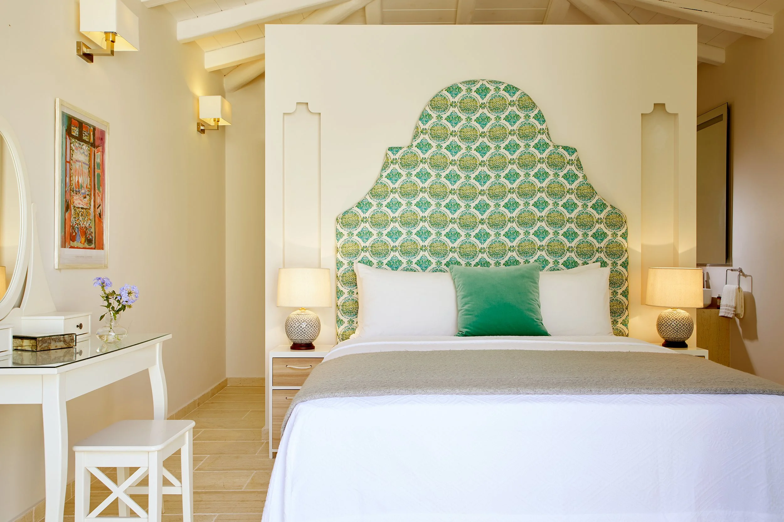A bedroom with a bed featuring a patterned green and blue headboard, white bedding, and a green pillow. There are two bedside tables with lamps, a white vanity with a mirror, a small table with a chair, a floral picture on the wall, and cream-colored walls with decorative wall sconces.