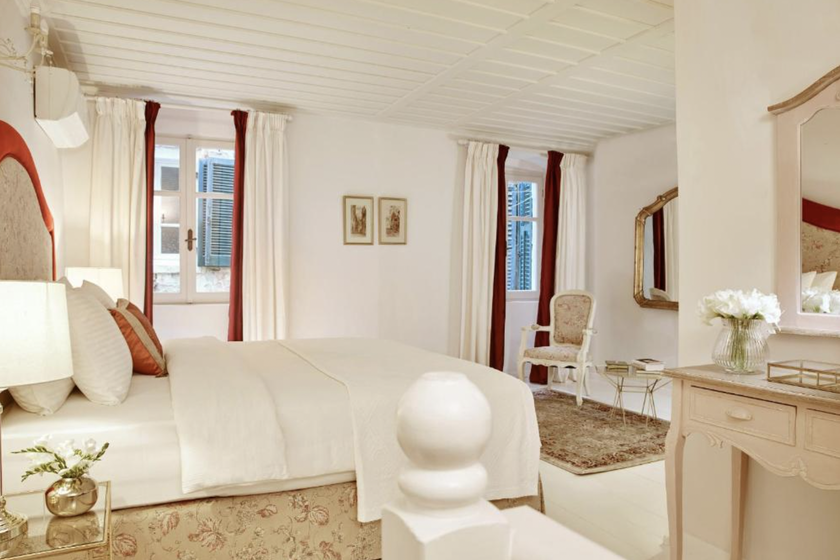 A cozy bedroom with a white bed, decorative pillows, cream and red curtains, a vintage armchair, a gold-framed mirror, and floral decor.