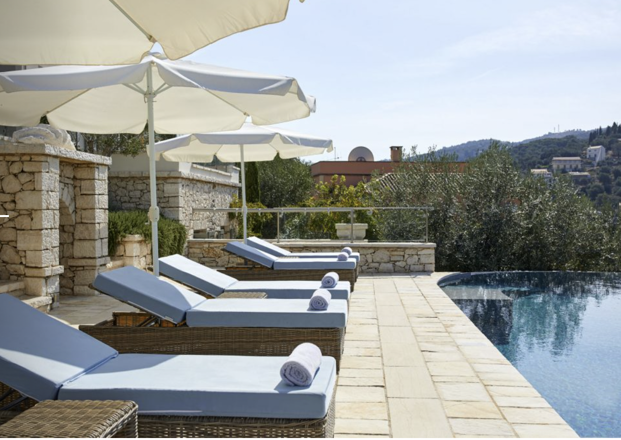 A luxury outdoor pool area with lounge chairs covered with blue cushions and rolled white towels, white umbrellas providing shade, stone walls, scenic hillside view in the background, clear sky, and calm pool water.