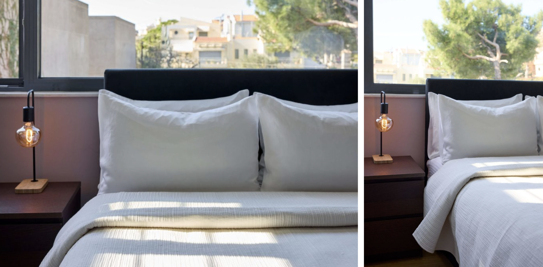 View of a neatly made bed with white bedding and pillows, flanked by a nightstand with a modern lamp, in front of a large window showing trees and buildings outside.