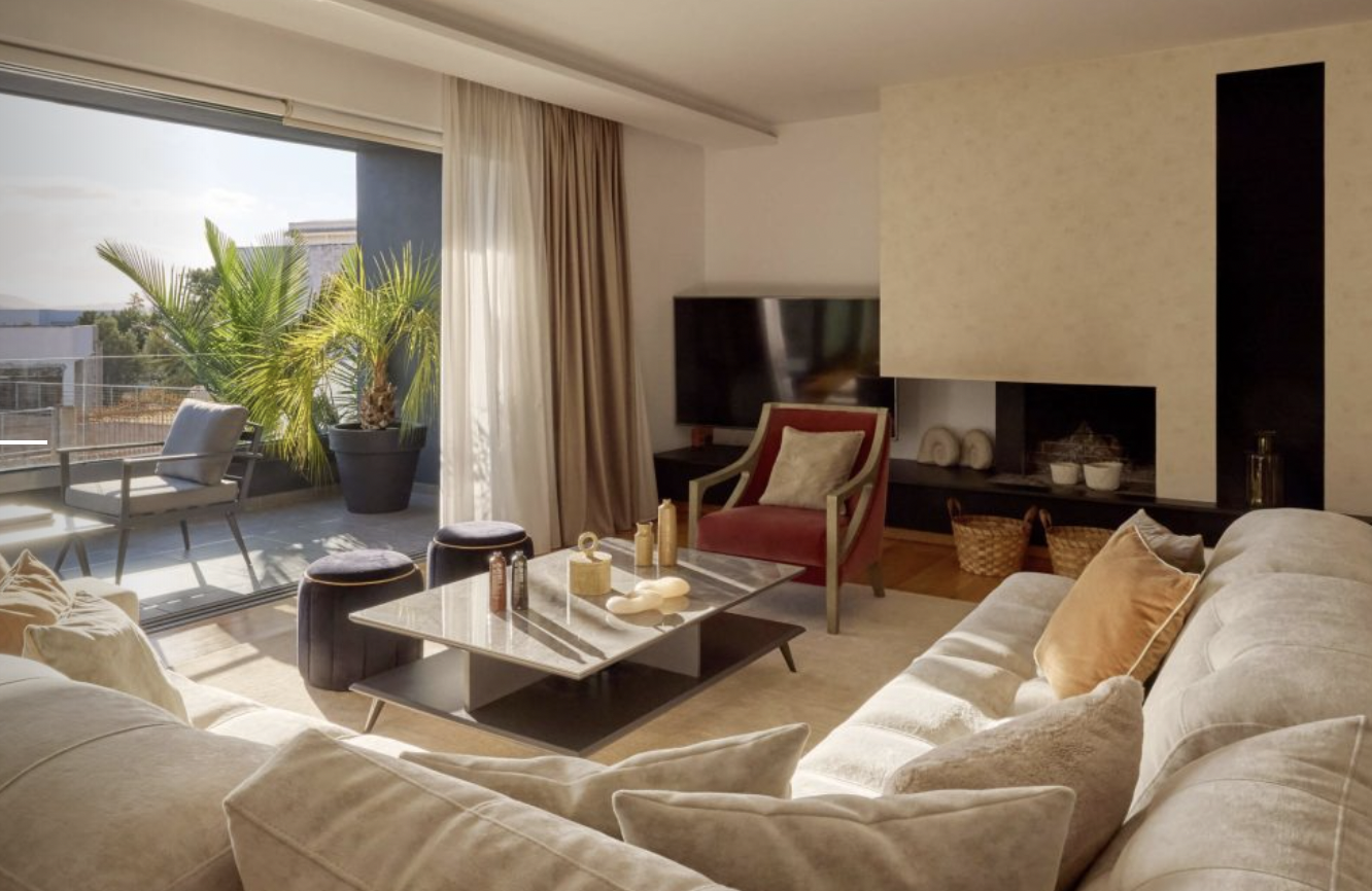 Bright living room with beige sofa, red armchair, coffee table, and large window with sliding glass door leading to a balcony with outdoor seating and a potted plant.
