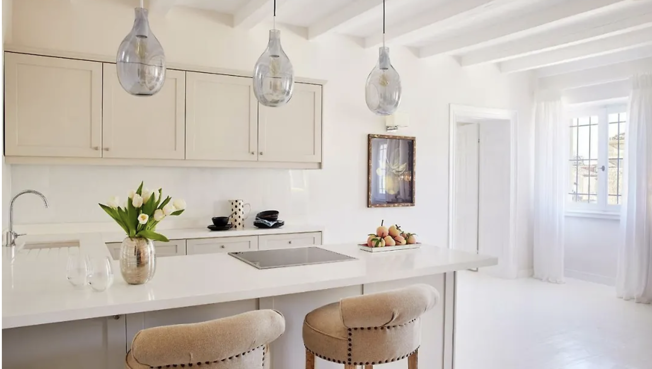 Bright, modern kitchen with white cabinets, a white island with bar stools, hanging light fixtures, a vase of white tulips, and a window with curtains letting in natural light.