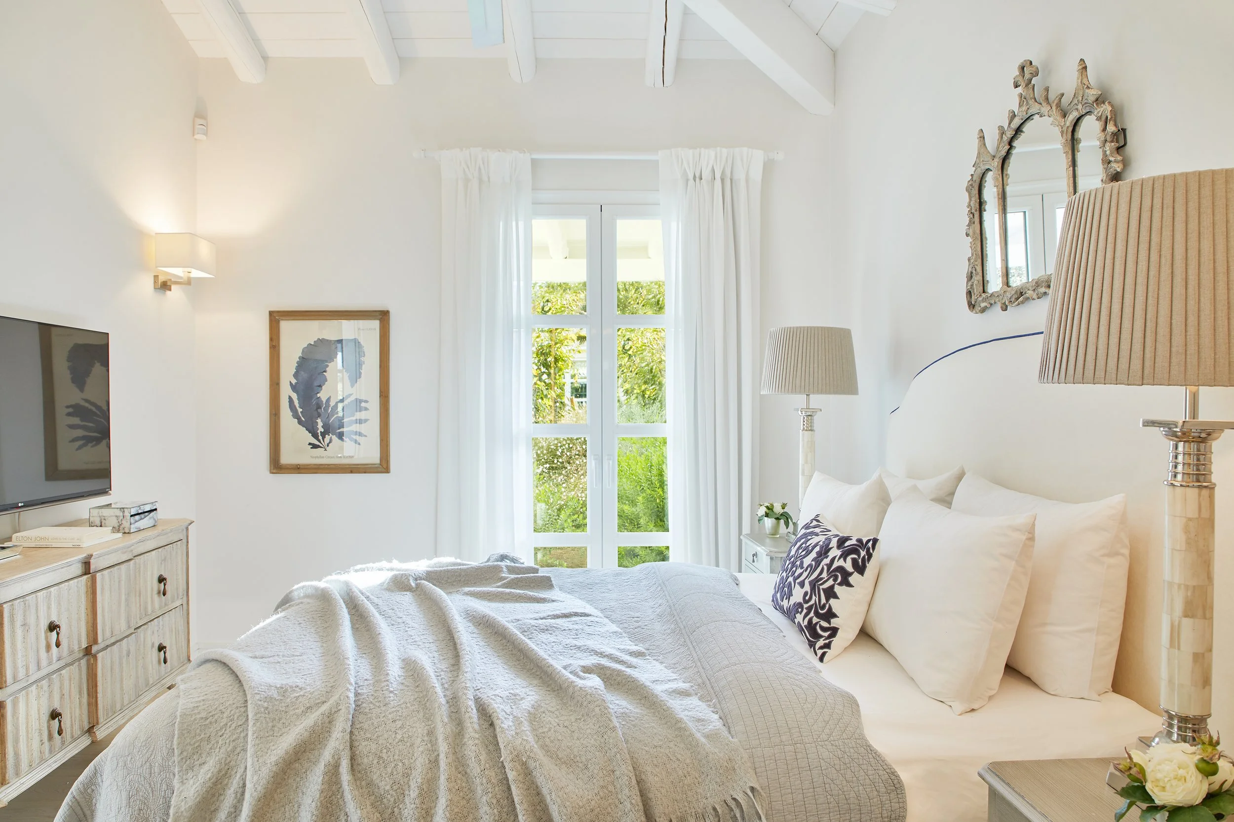 Bright bedroom with a large white bed, beige nightstand, table lamps, white curtains, and a window showing greenery outside. White walls and ceiling beams add to the airy feel.