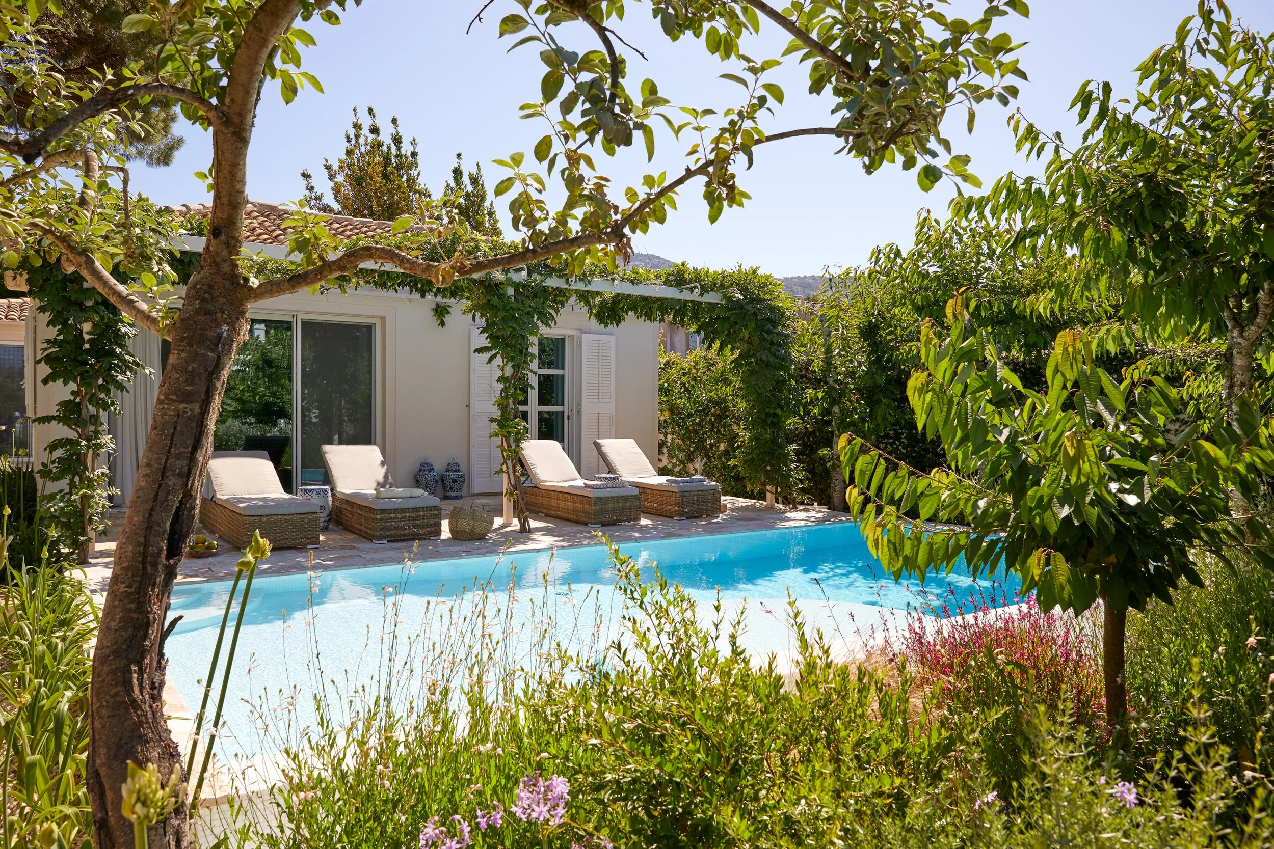 A backyard pool area with four lounge chairs, surrounded by lush trees and plants, in front of a white house with sliding glass door and window with shutters.