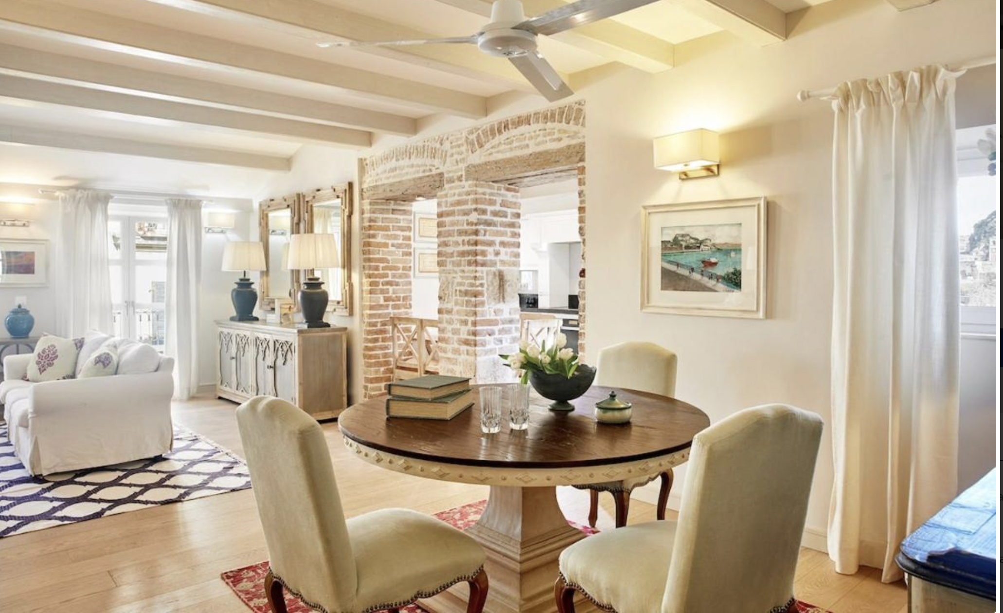 Bright living and dining area with a round wooden table and four cream upholstered chairs. The table has a black bowl with white flowers, books, and glassware. In the background, there is a white sofa with floral pillows, a wooden sideboard with blue vases, brick and white walls, window with white curtains, and a ceiling fan.