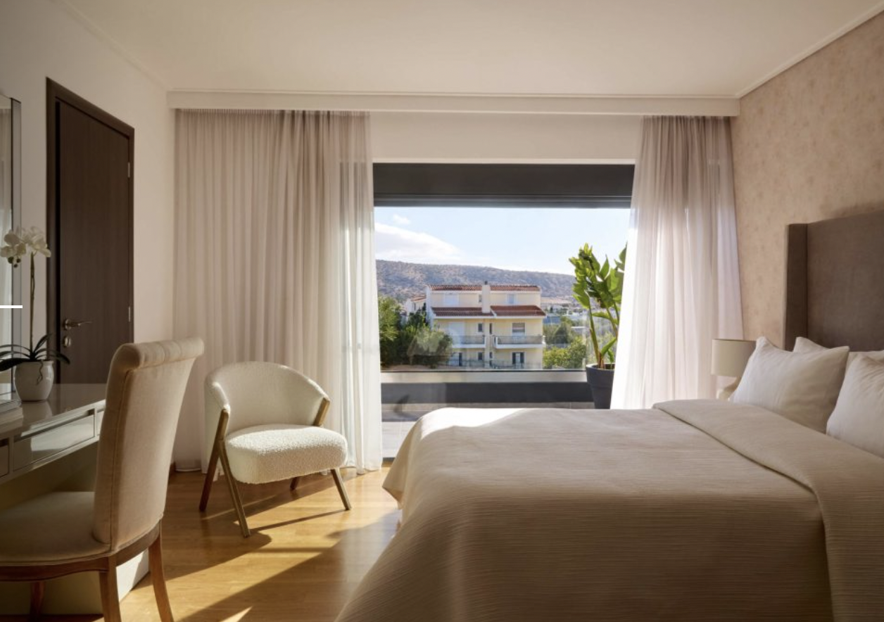 Hotel room with a large bed, a desk with a chair, a white armchair, a potted plant, and a sliding glass door with white curtains leading to a balcony, showing a view of nearby buildings and distant mountains.