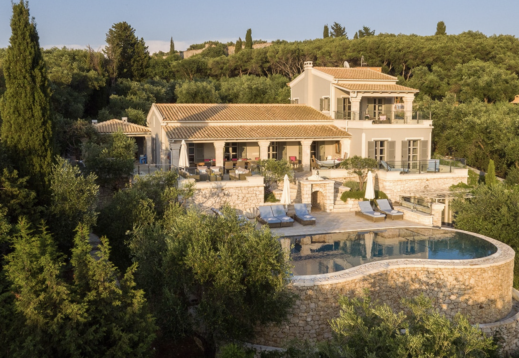 Large Mediterranean-style house with beige walls and terracotta roof, surrounded by lush greenery, with a stone patio, pool, and outdoor furniture.