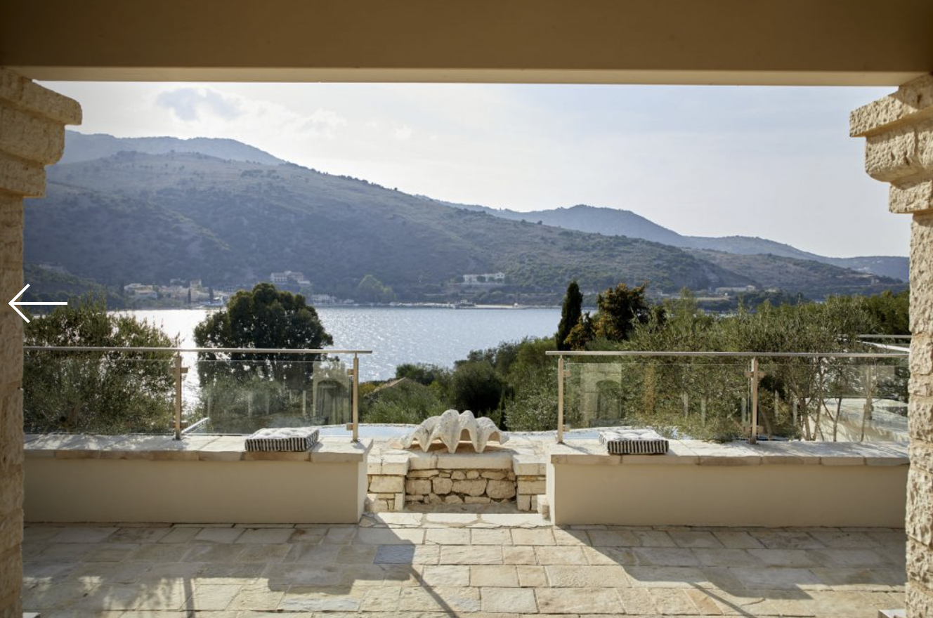 View of a lake surrounded by hills, seen from a terrace with stone flooring and glass railing, with decorative shells and small pillows on the ledge.
