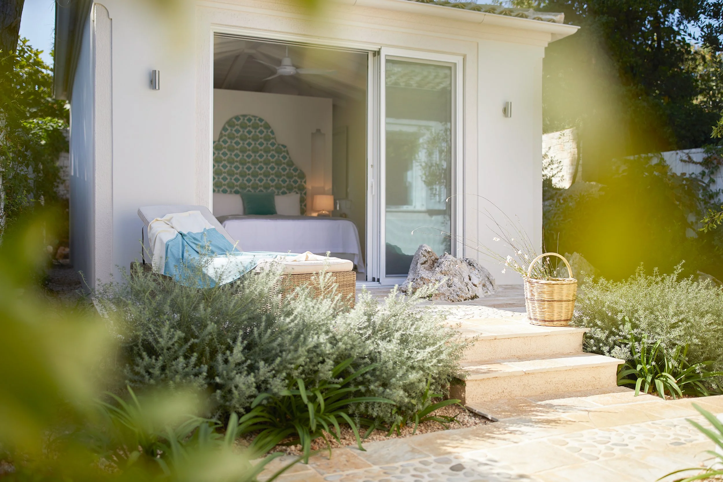 A view of a backyard with steps leading to a sliding glass door, opening into a bedroom with a bed visible inside. There is a lounge chair with a towel draped over it, and a wicker basket on the patio. The yard is surrounded by green shrubs and plants, with sunlight illuminating the scene.