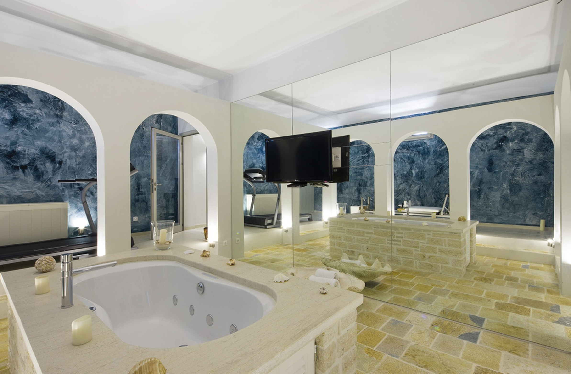 A luxurious bathroom with a large bathtub, candle holders, a wall-mounted TV, a treadmill, and blue textured walls, all reflected in a mirrored wall.
