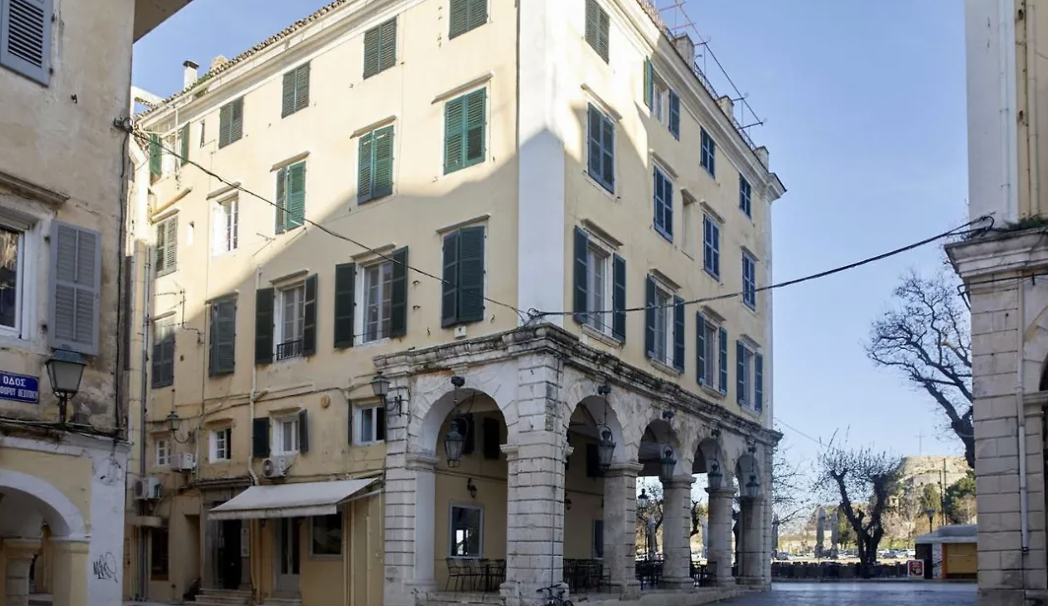A multi-story yellow building with green shutters and arched entrance supported by columns, situated on a street corner with leafless trees and a clear blue sky.