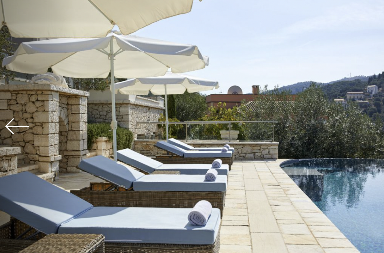 Swimming pool area with lounge chairs, rolled towels, and umbrellas, overlooking a hillside with trees and buildings in the background.
