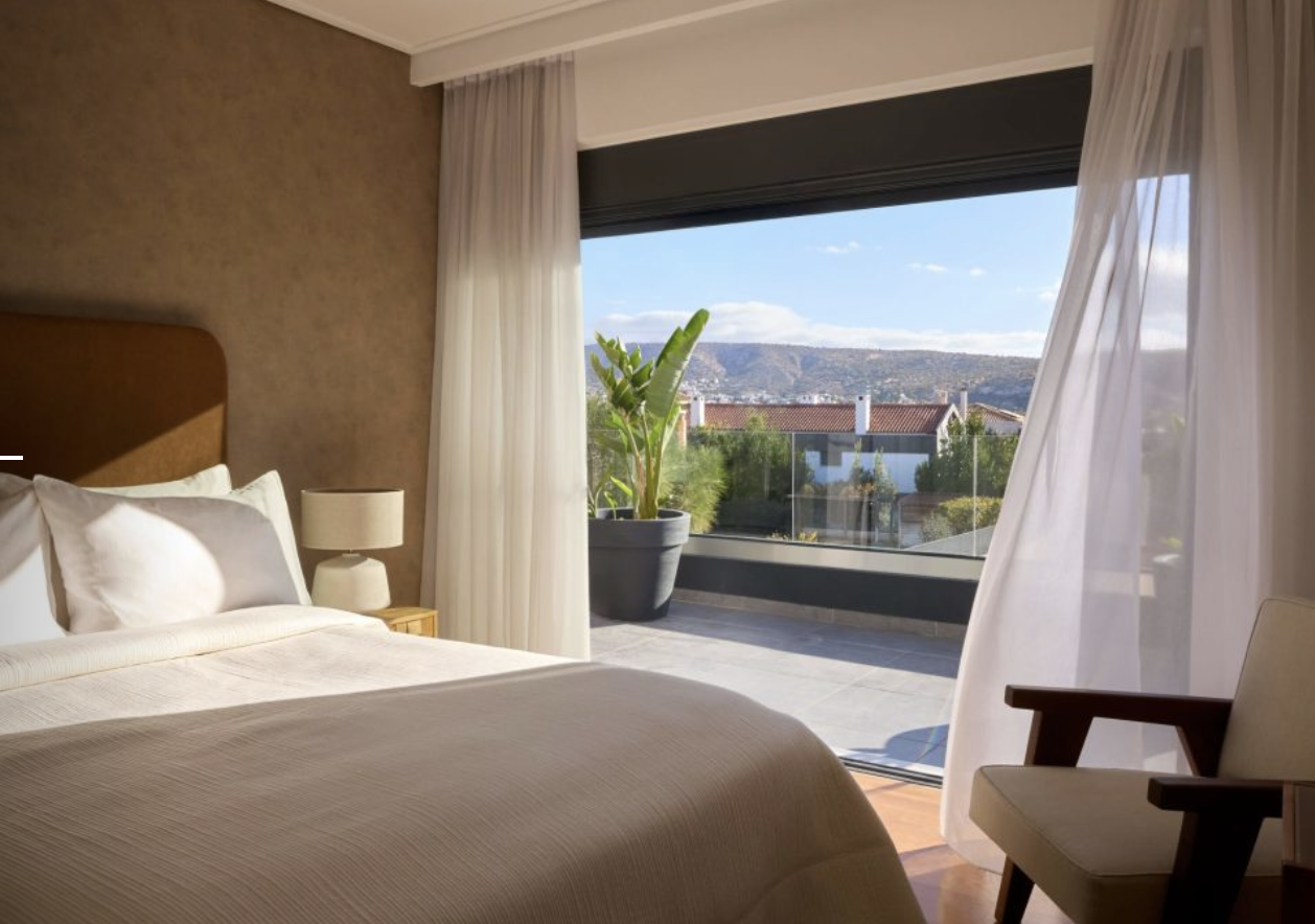 A bedroom with a large window overlooking a balcony with a potted plant, neighboring houses, and hills in the distance. The bed is made with white sheets and pillows, and there is a bedside table with a lamp. Sheer curtains frame the window.