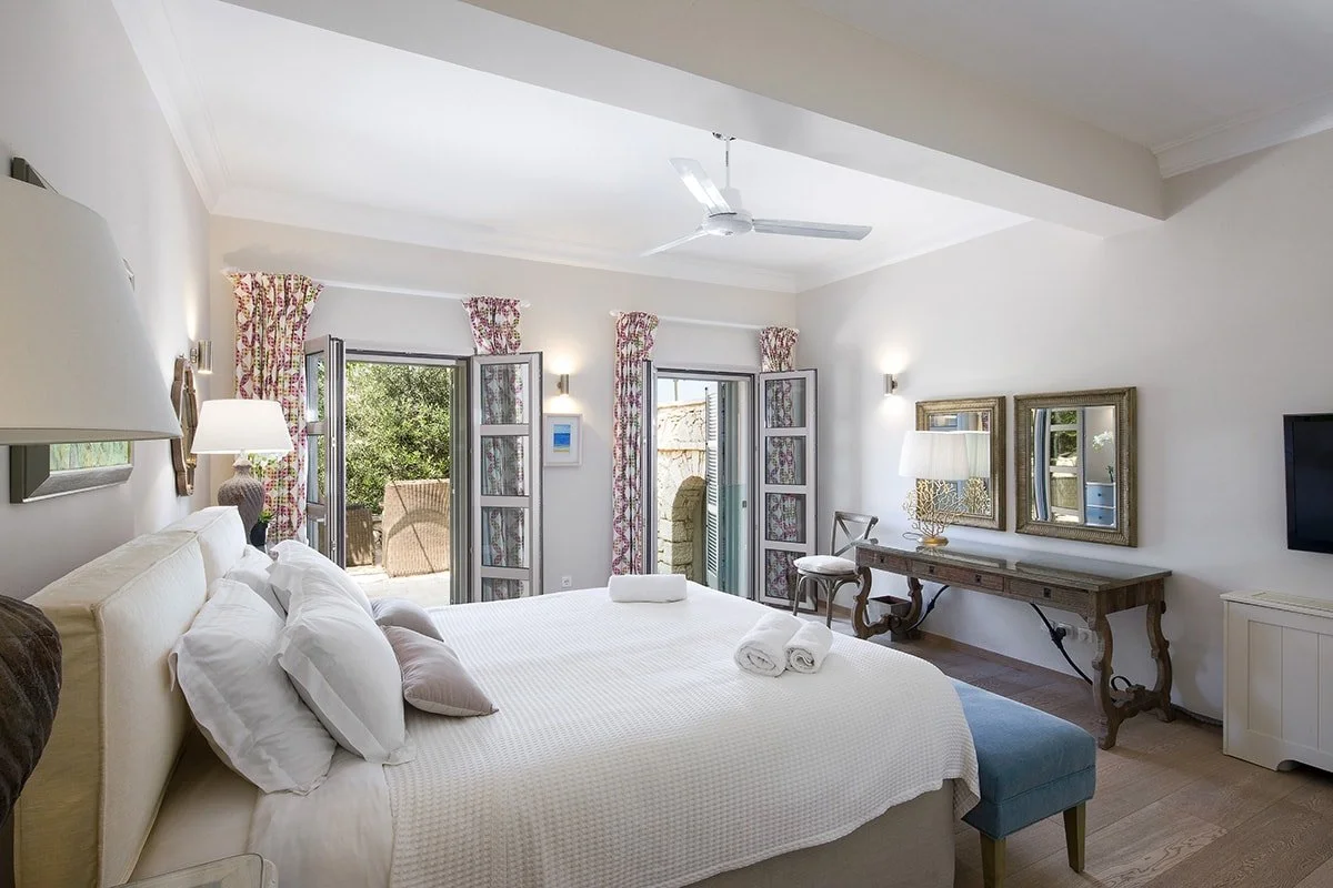 Bright hotel bedroom with a large bed, two open windows with floral curtains, and outdoor seating visible outside. Decor includes lamps, framed mirrors, a wooden desk, and a flat-screen TV.