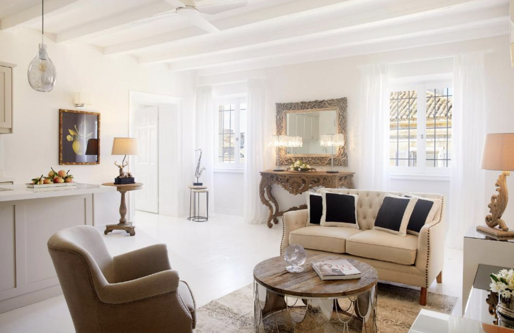 Bright living room with white walls, ceiling, and floors, featuring a beige sofa with black and beige pillows, a round wooden coffee table with decorative items, an armchair, lamps, art on the walls, and windows with white curtains.