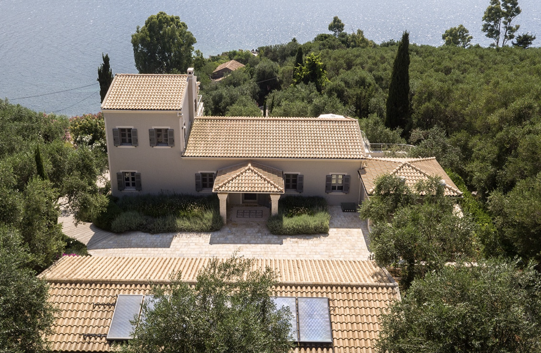 Aerial view of a large house with a tiled roof surrounded by trees and greenery near a body of water.
