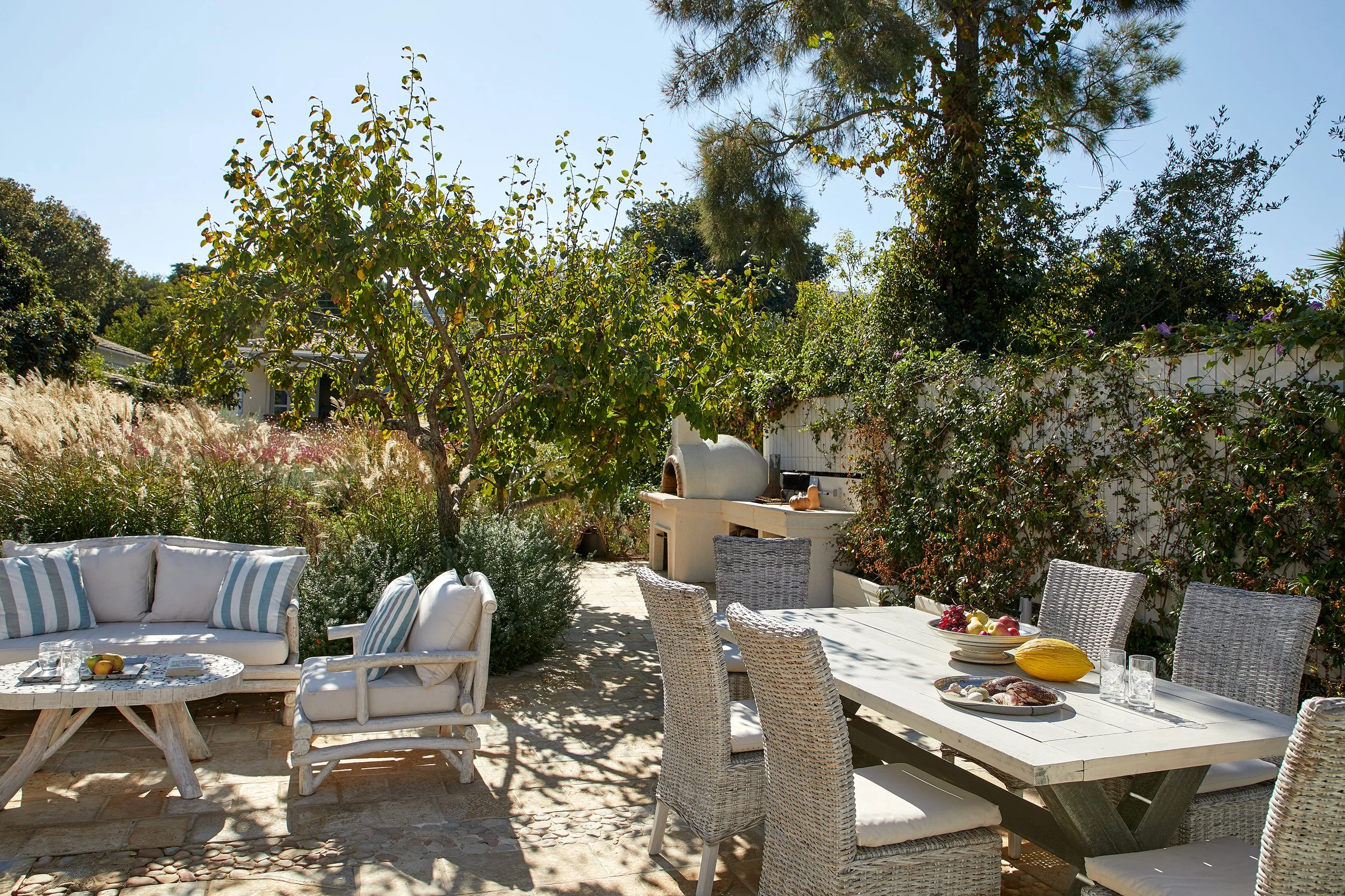 Outdoor patio with white cushioned furniture, a dining table with food and glasses, trees, plants, and a built-in pizza oven.