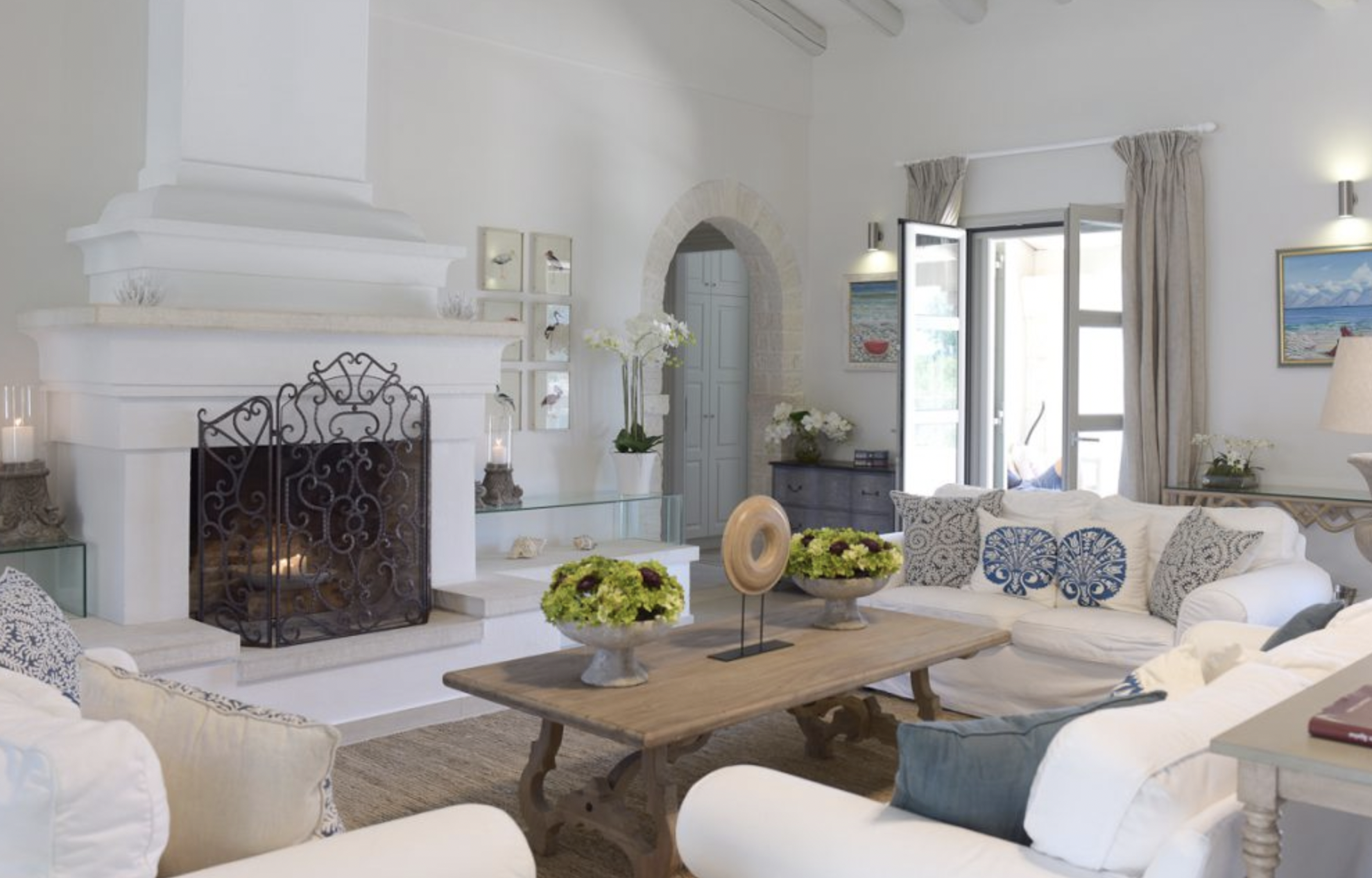 A living room with white walls, a white stone fireplace, and cream-colored sofas. There are decorative pillows, vases of flowers, and artwork on the walls. A window with open curtains lets in natural light.