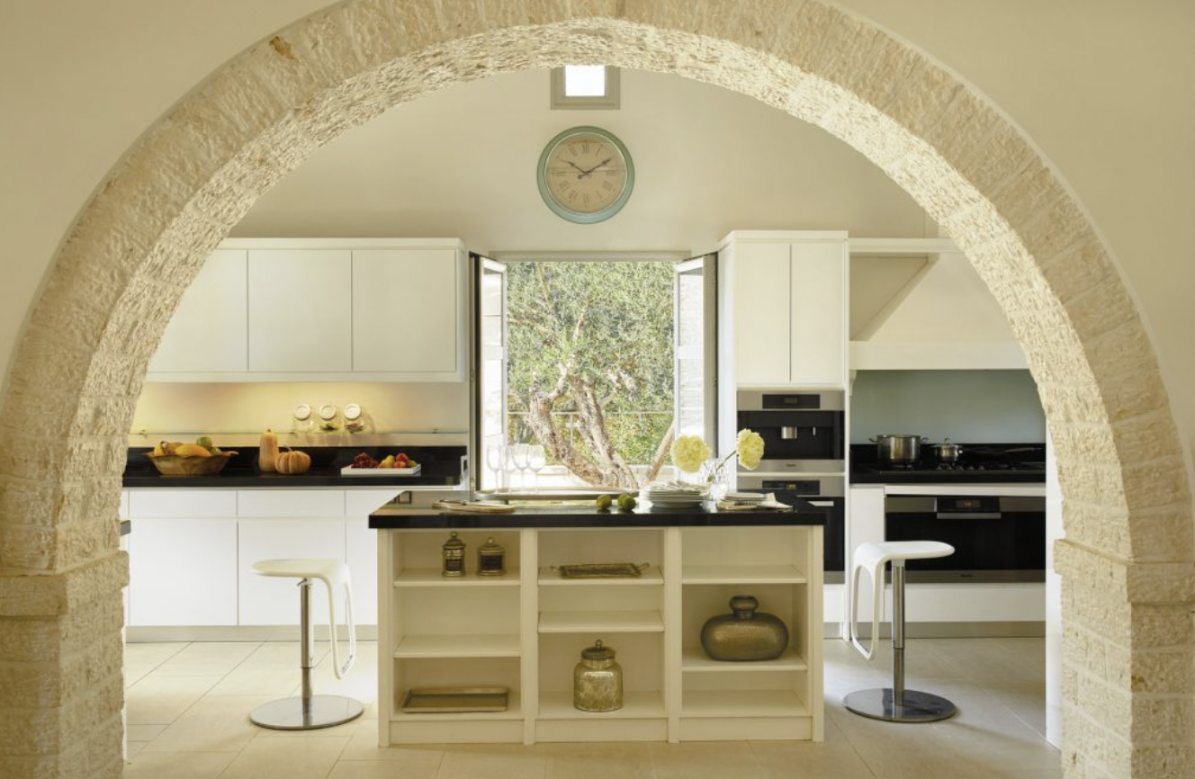 Modern kitchen with white cabinets, black countertops, and a large window showing a tree outside, with a stone arch framing the view.