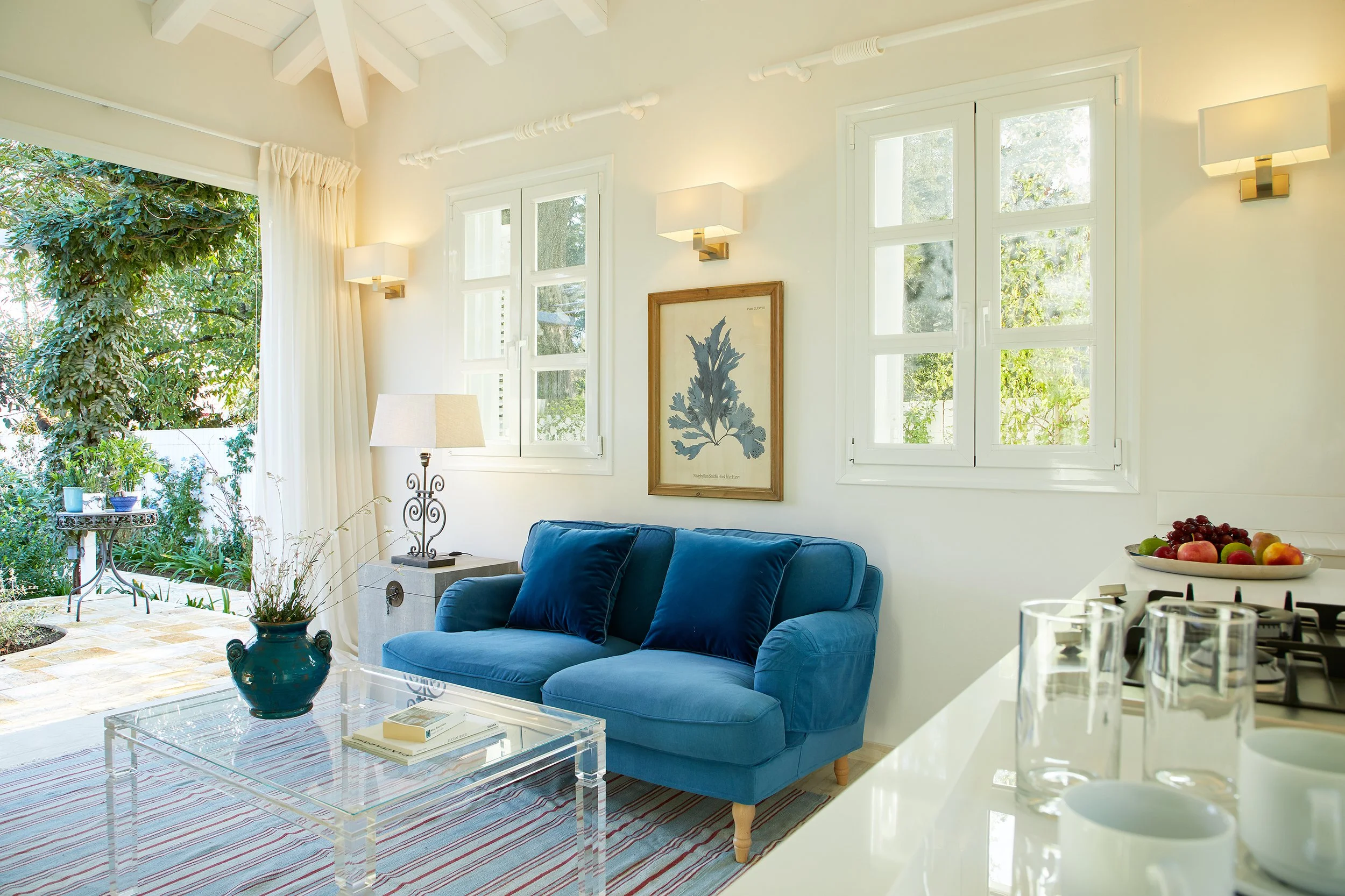 Bright living room with white walls, multiple windows, and a blue sofa. Decor includes a framed botanical print, wall-mounted lamps, a table lamp, and a vase with dried grass on a clear coffee table. The room opens to a patio with garden greenery visible outside.