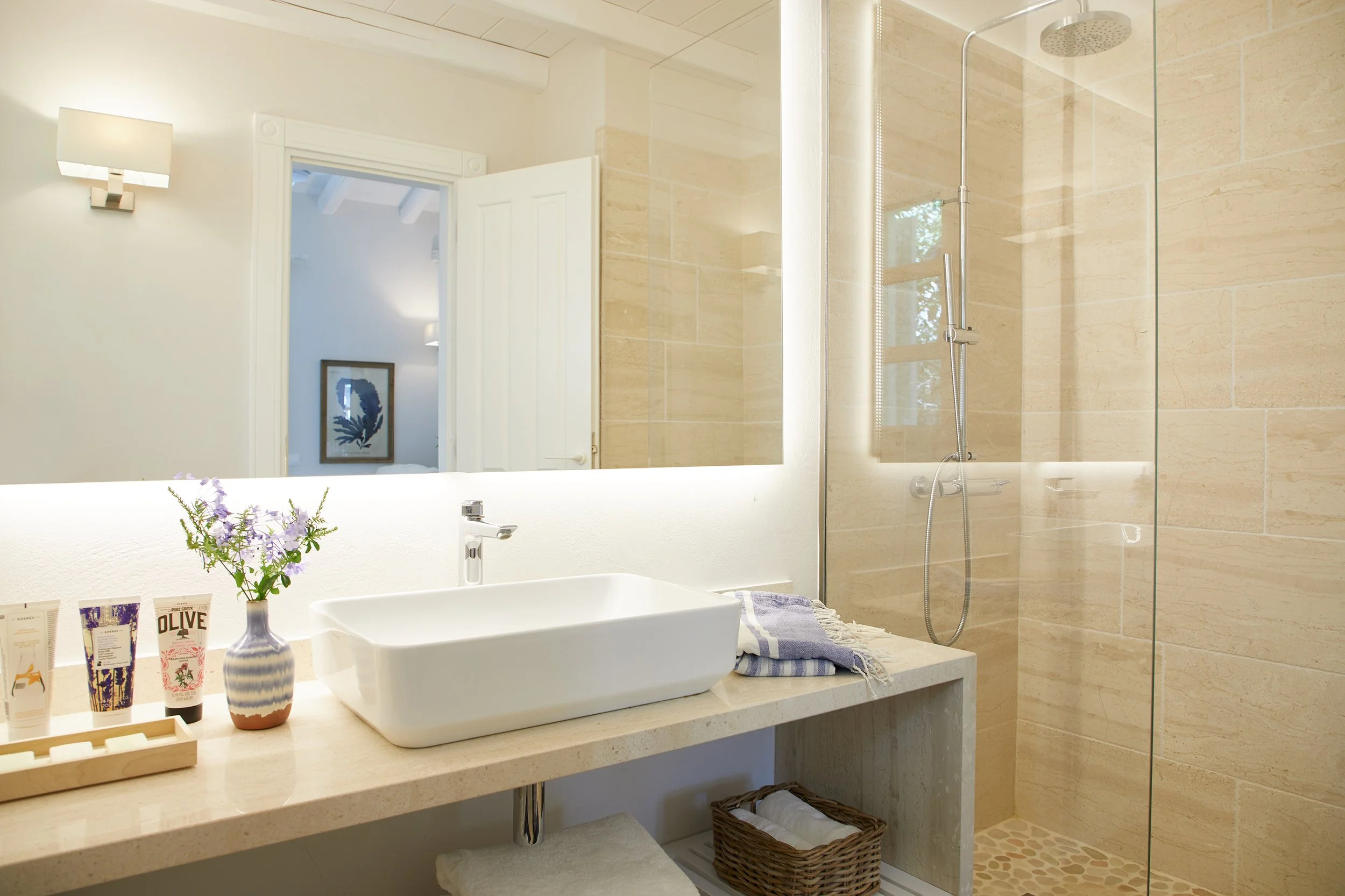 Modern bathroom with a large mirror, white vessel sink, beige countertops, and a walk-in shower with a glass door and beige stone tiles.