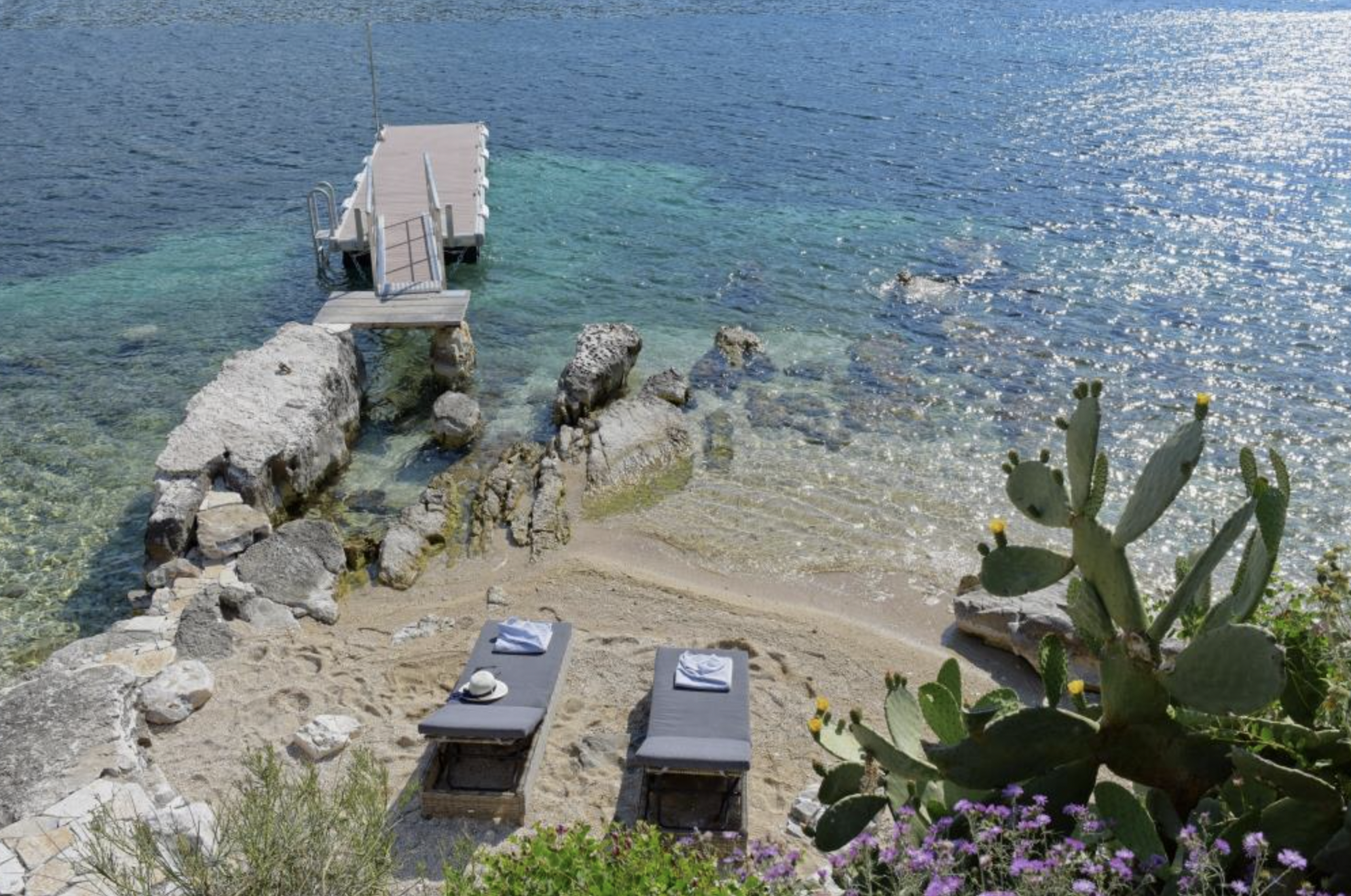 Two lounge chairs on a sandy beach with rolled towels and a hat, overlooking clear blue ocean water, with a cactus and purple flowers in the foreground and a small pier extending into the water.