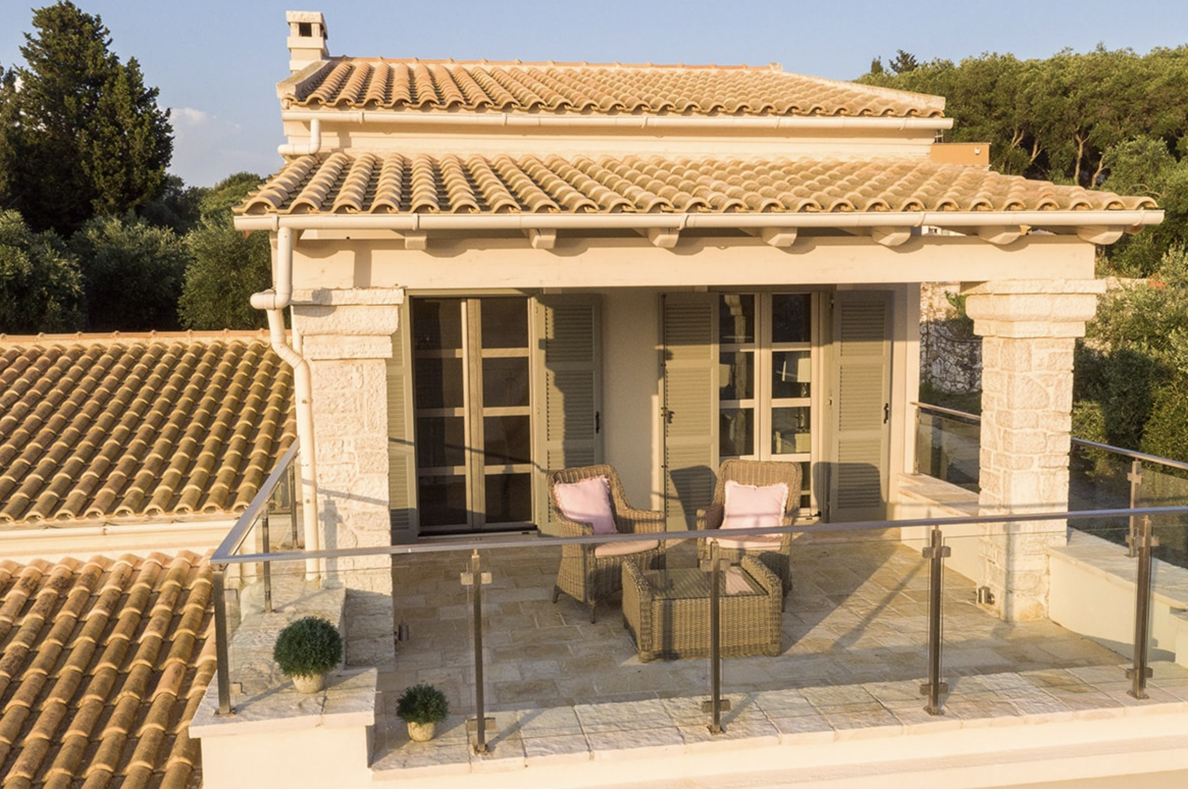 A rooftop terrace with wicker furniture, pink pillows, and a glass railing, overlooking a building with stone columns and a tiled roof, surrounded by greenery.