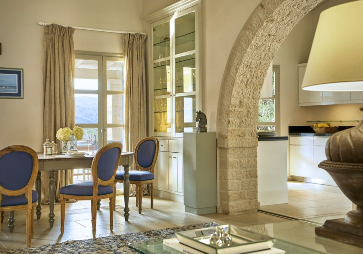 Cozy dining room with wooden table, blue upholstered chairs, glass cabinet, large window with beige curtains, stone archway leading to kitchen, and decorative items.