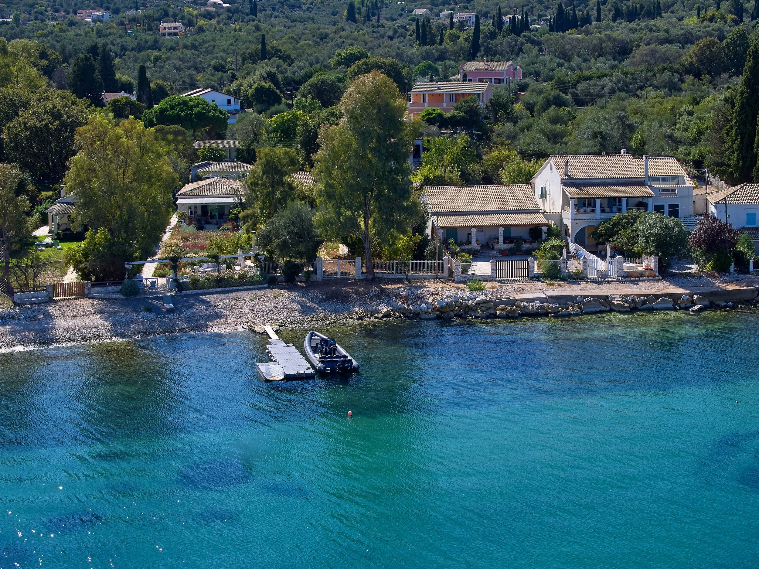 Aerial view of waterfront houses on a hillside with trees, rocky shoreline, and a floating dock with a small boat, surrounded by clear blue water.