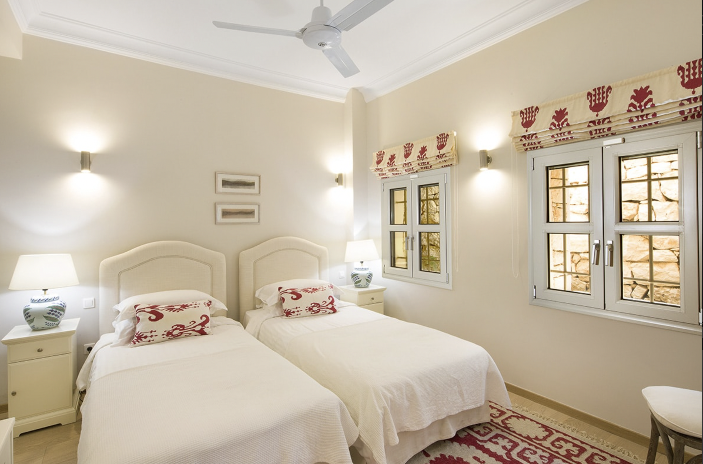 A bedroom with two twin beds with white linens and decorative pillows, two white nightstands with lamps, two windows with patterned valances, beige walls, framed artwork, and a ceiling fan.