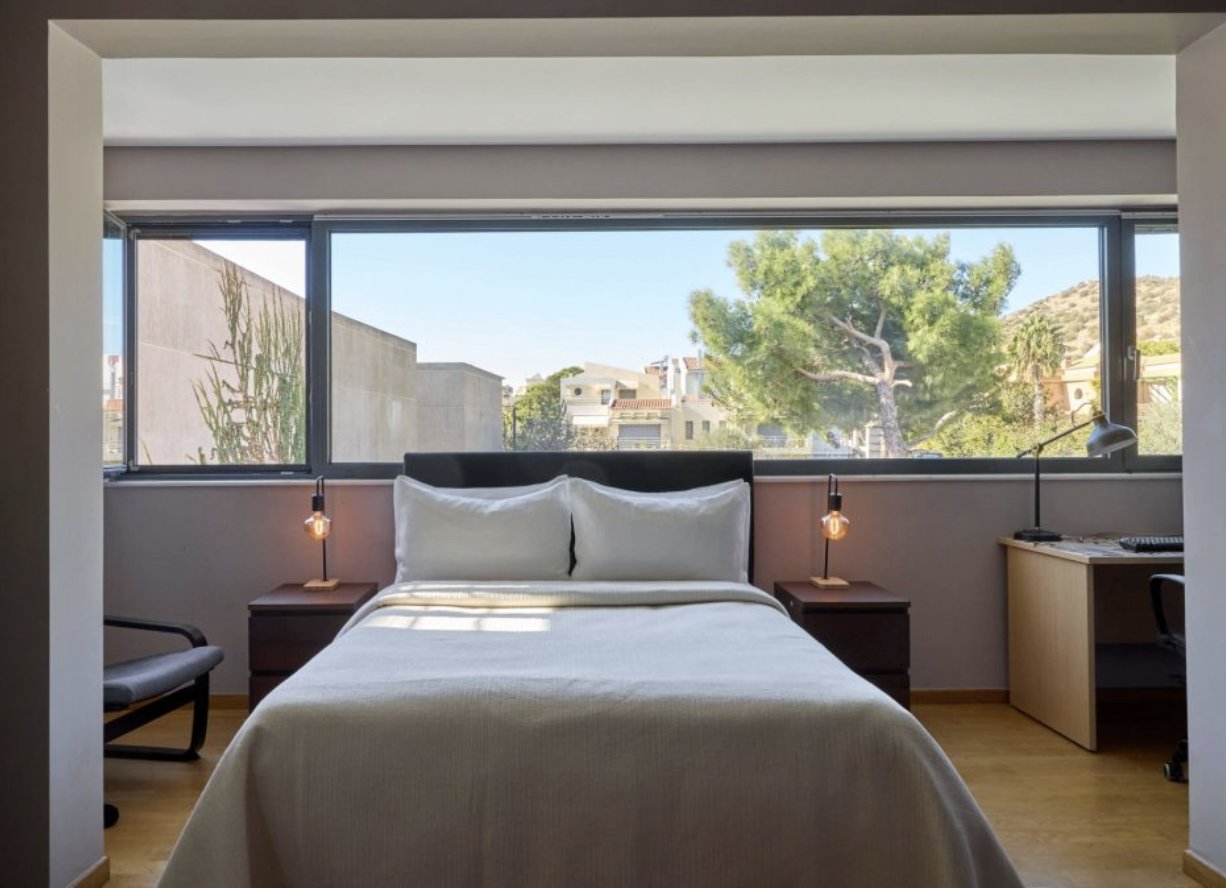 A modern bedroom with a large window, a bed with white bedding, two small nightstands with lamps, a chair, and a desk with a lamp and computer, overlooking trees and buildings outside.