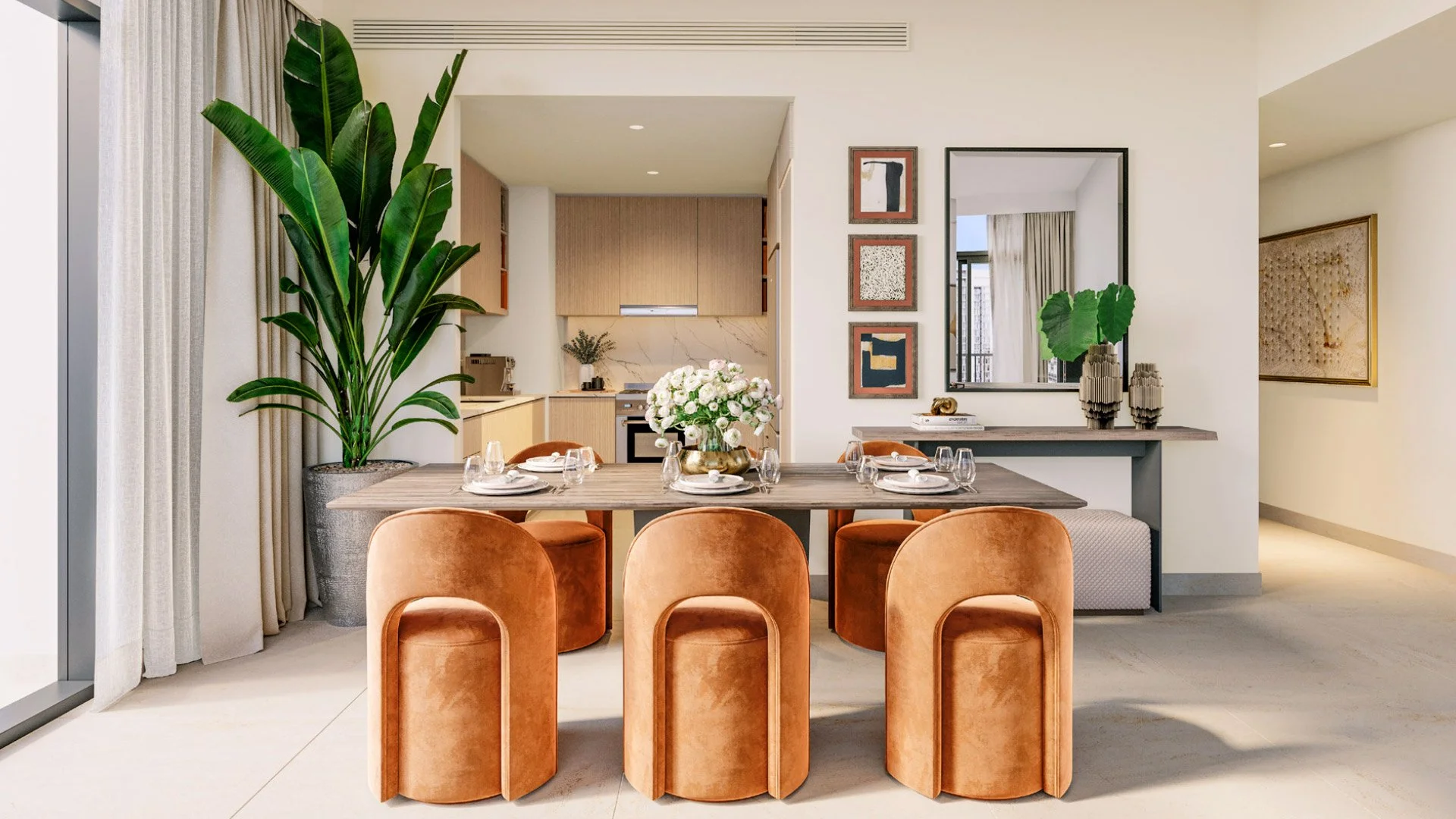 Modern dining room with a wooden table set for six, surrounded by coral velvet chairs, and decorated with a vase of white flowers. Large green plant in a gray pot on the left, and art pieces on the wall. Open kitchen with beige cabinets in the backgr