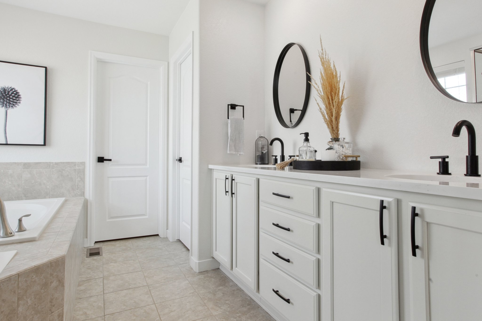 After the Flip: bathroom styled with white cabinetry, black hardware and faucets, beige tiled flooring, and oval mirrors. The space is finished with simple decorative styling, including dried plants, creating a clean, balanced look designed to improv
