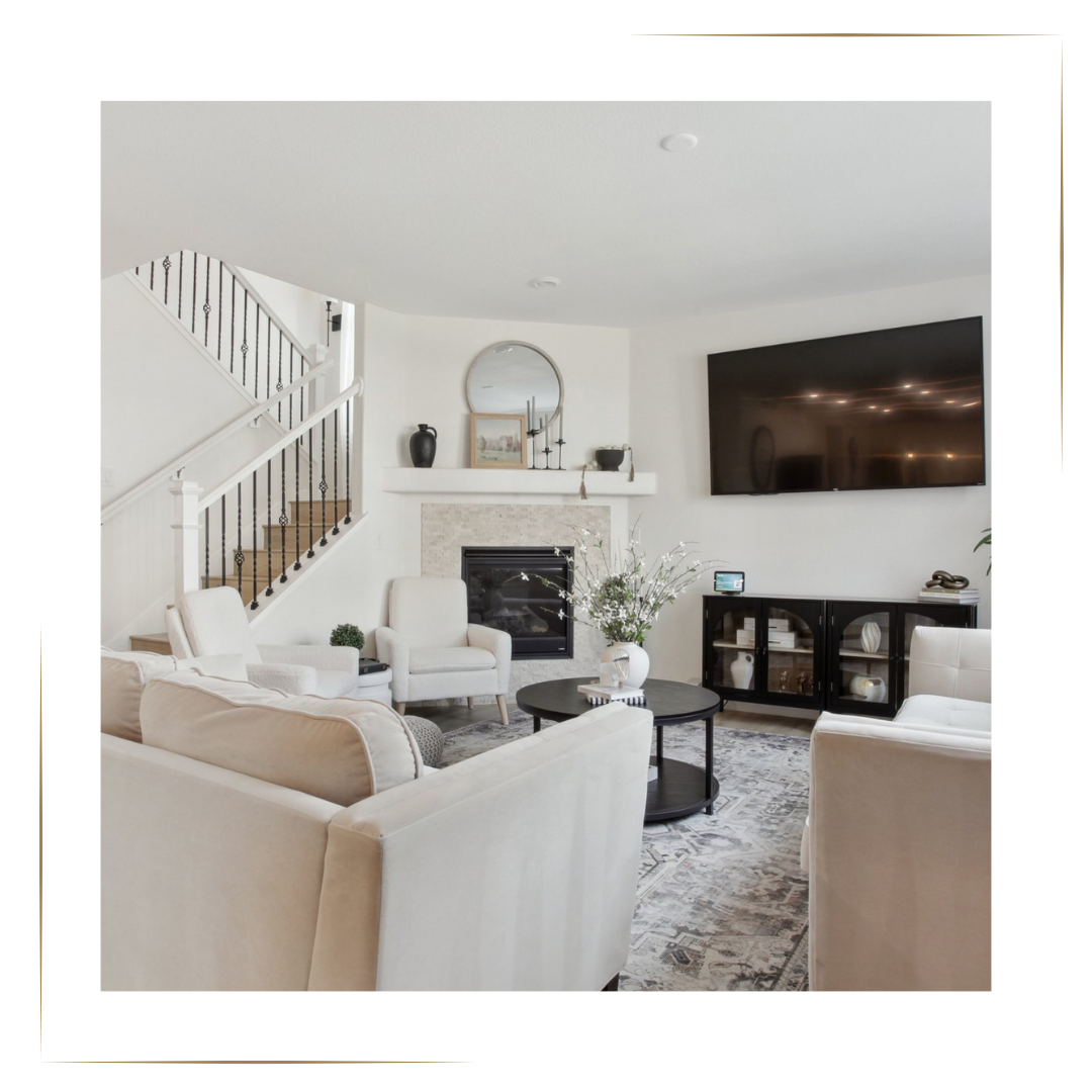 Home Staging Harmony Homes by Amanda Modern living room with white sofas, armchairs, a black coffee table with a vase of flowers, a TV mounted on the wall, a fireplace, and a staircase with black railing in the background.