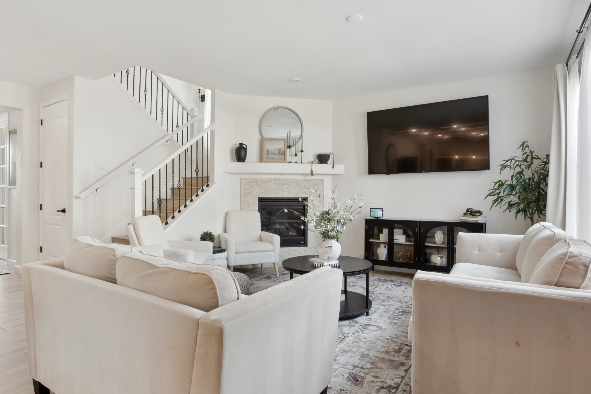 Living room after Flip Design and staging, featuring cream sofas, a black coffee table, a fireplace with a round mirror, a staircase, and a large window with white curtains.