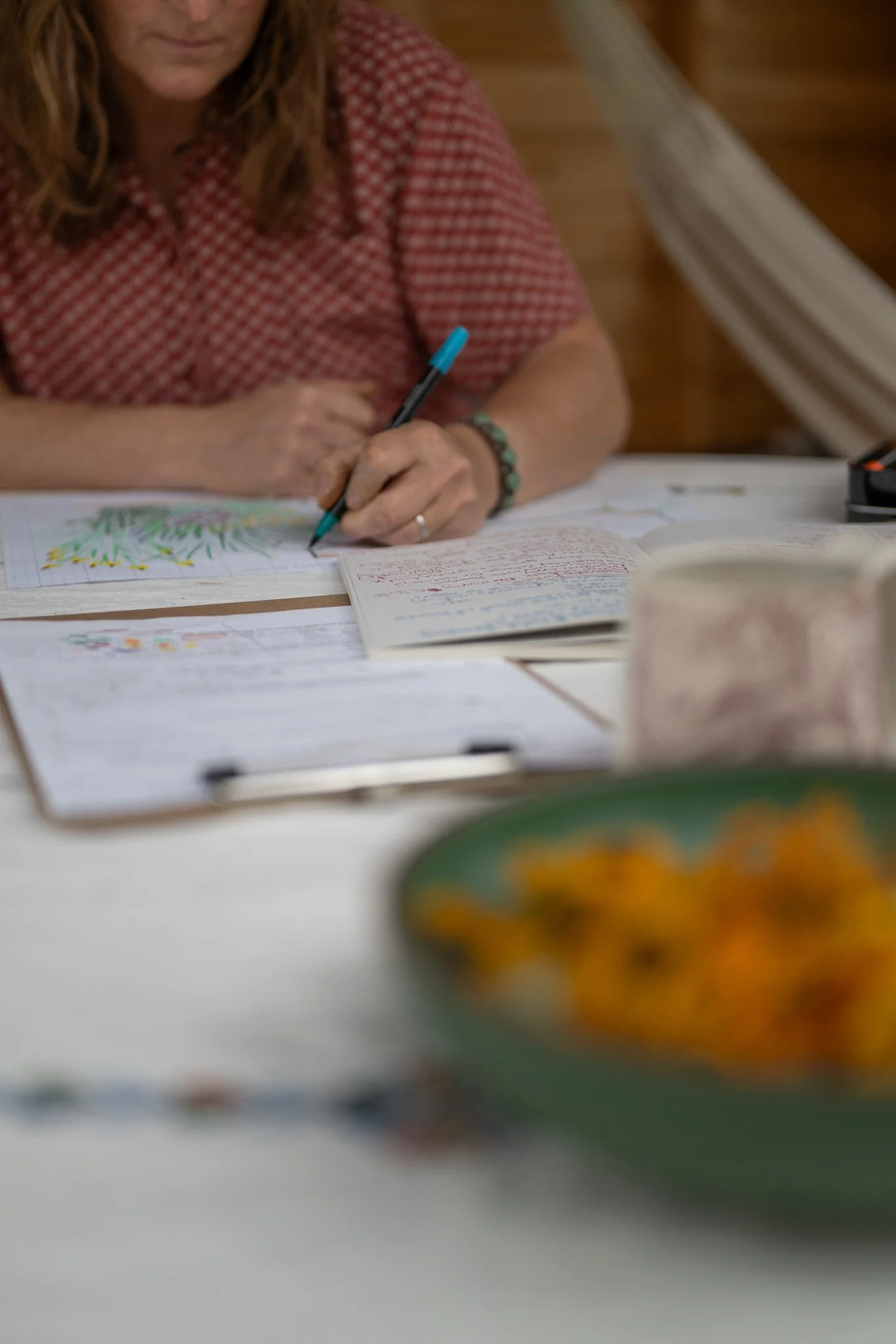 A woman in a red checkered shirt sitting at a table, writing or drawing with a blue pen, surrounded by notebooks, papers, and a mug.