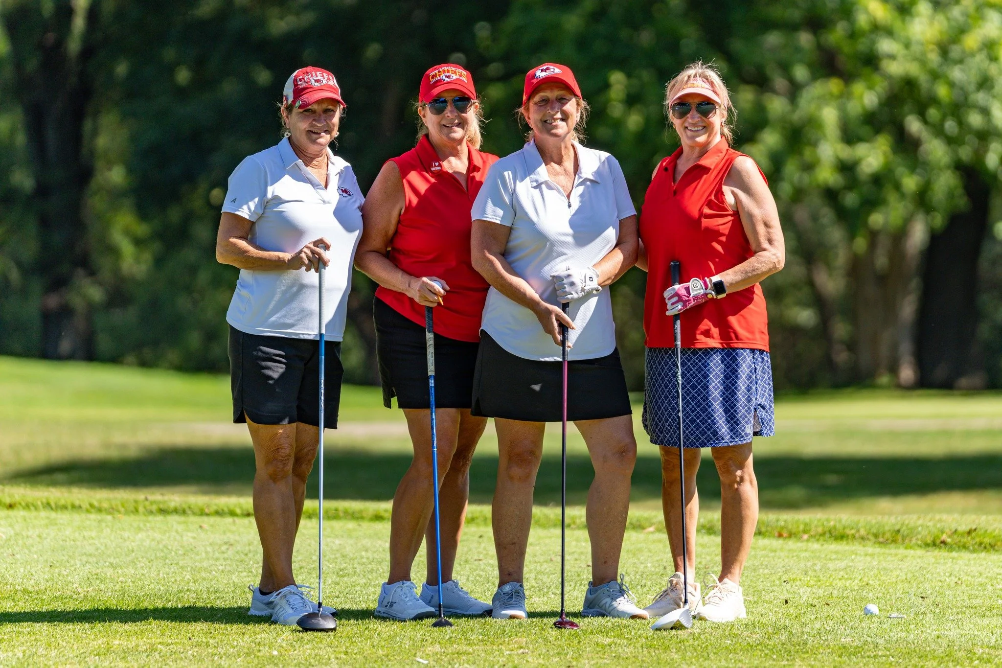 Four women standing on a golf course holding golf clubs, dressed in golf attire and hats, with trees and green grass in the background.