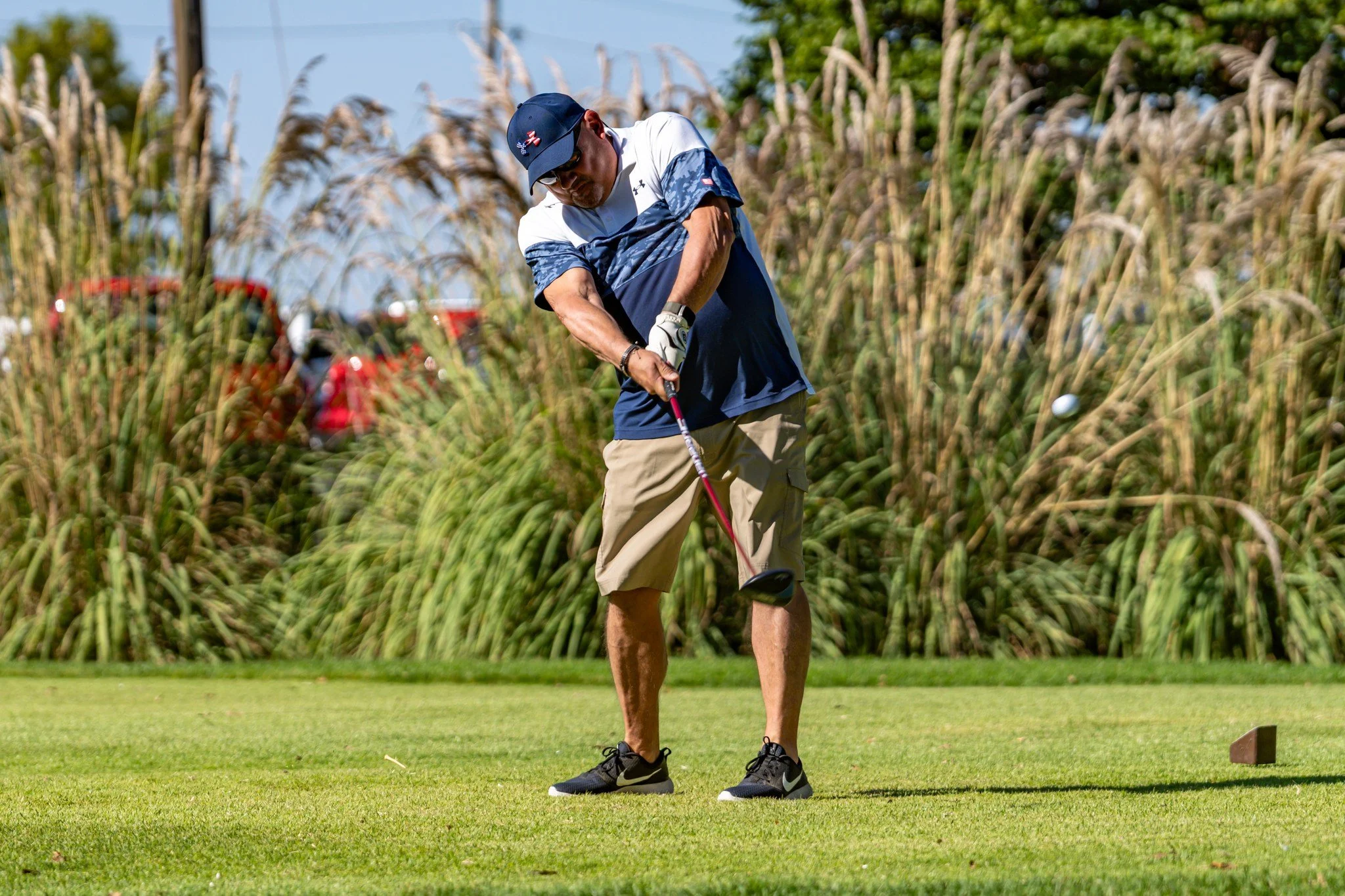 A man playing golf on a lush green fairway, preparing to strike the golf ball with a club, surrounded by tall grass and trees.
