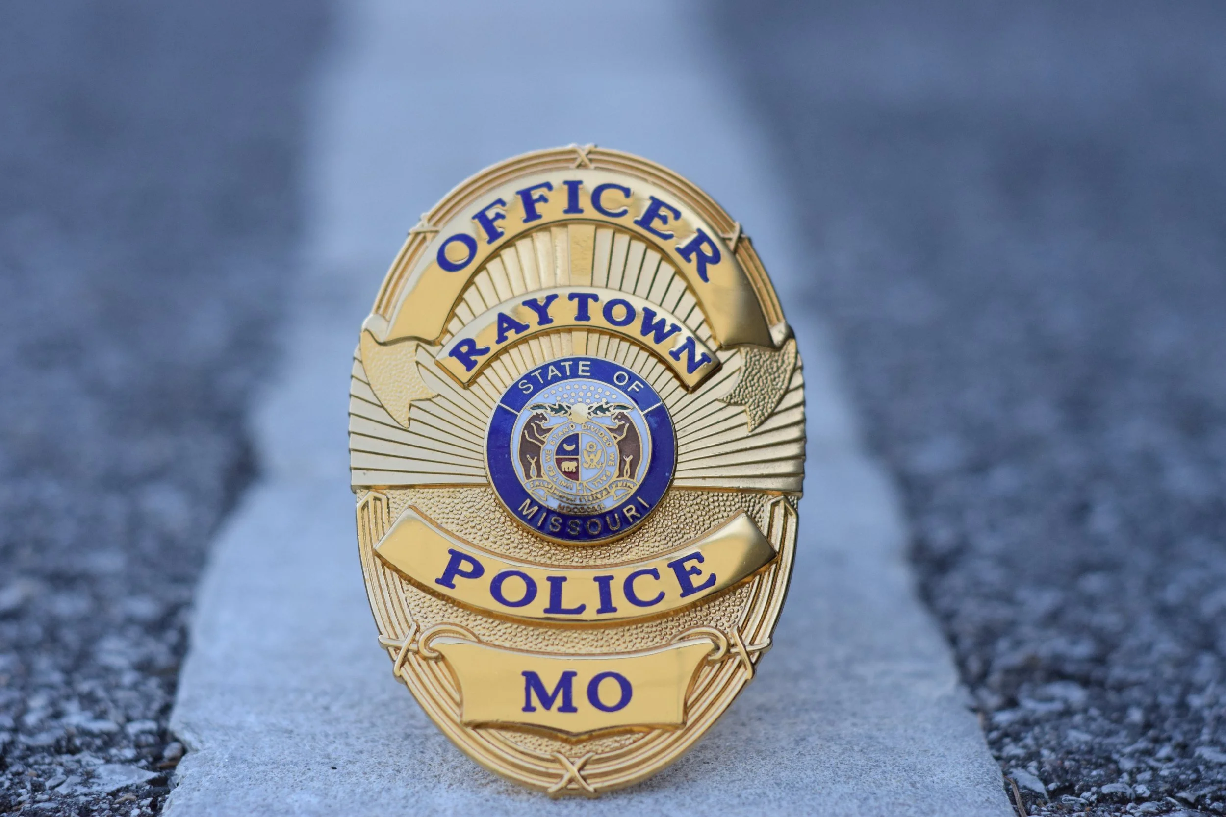 A Missouri police badge from Raytown Office, depicting a circle with the state emblem in the center and gold rays radiating outward, placed on a concrete surface.