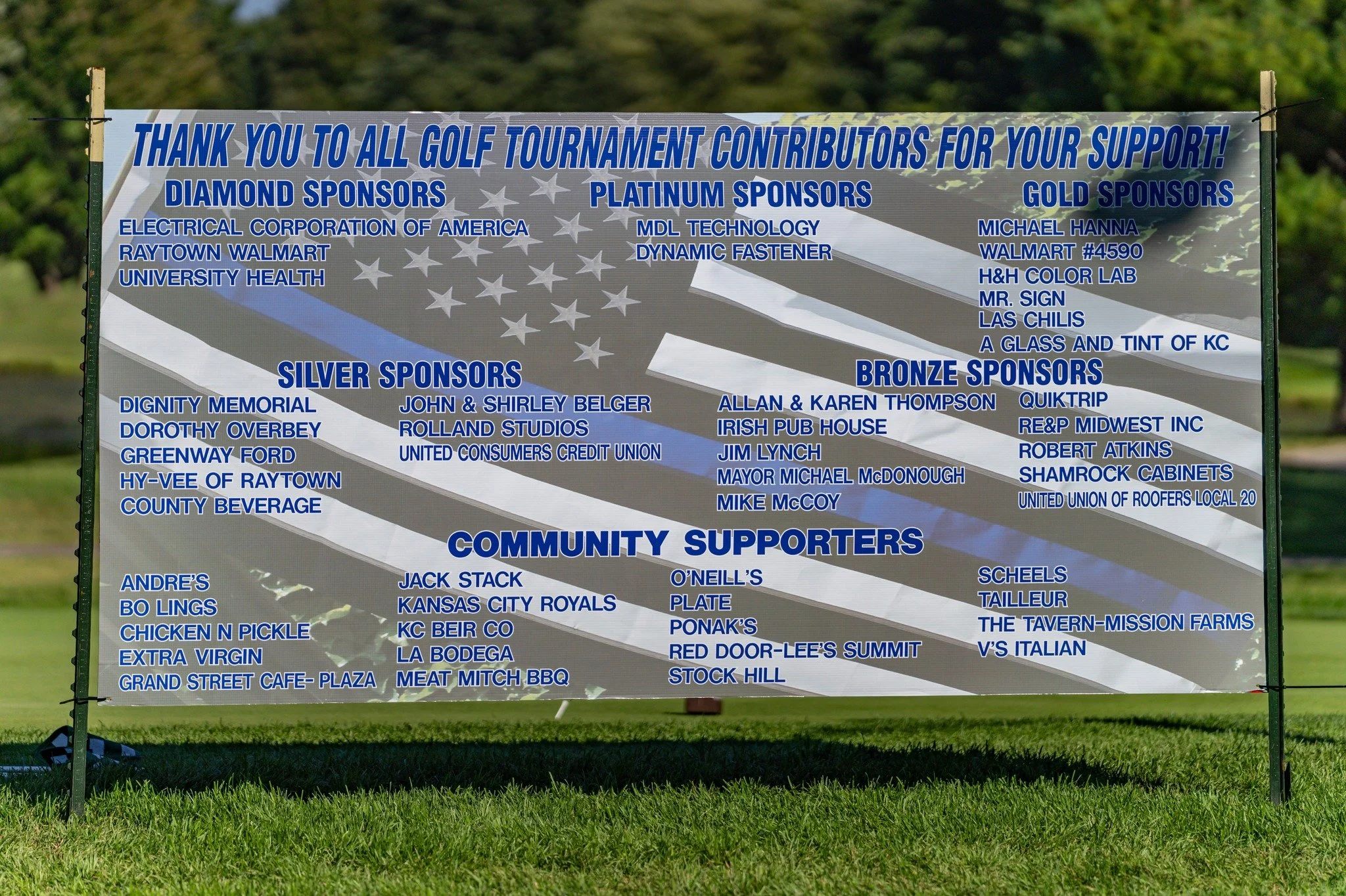A large outdoor banner thanking golf tournament contributors, with categories for diamond, platinum, gold, silver, bronze sponsors, and community supporters, displaying names of various sponsors and supporters on a background of the American flag.