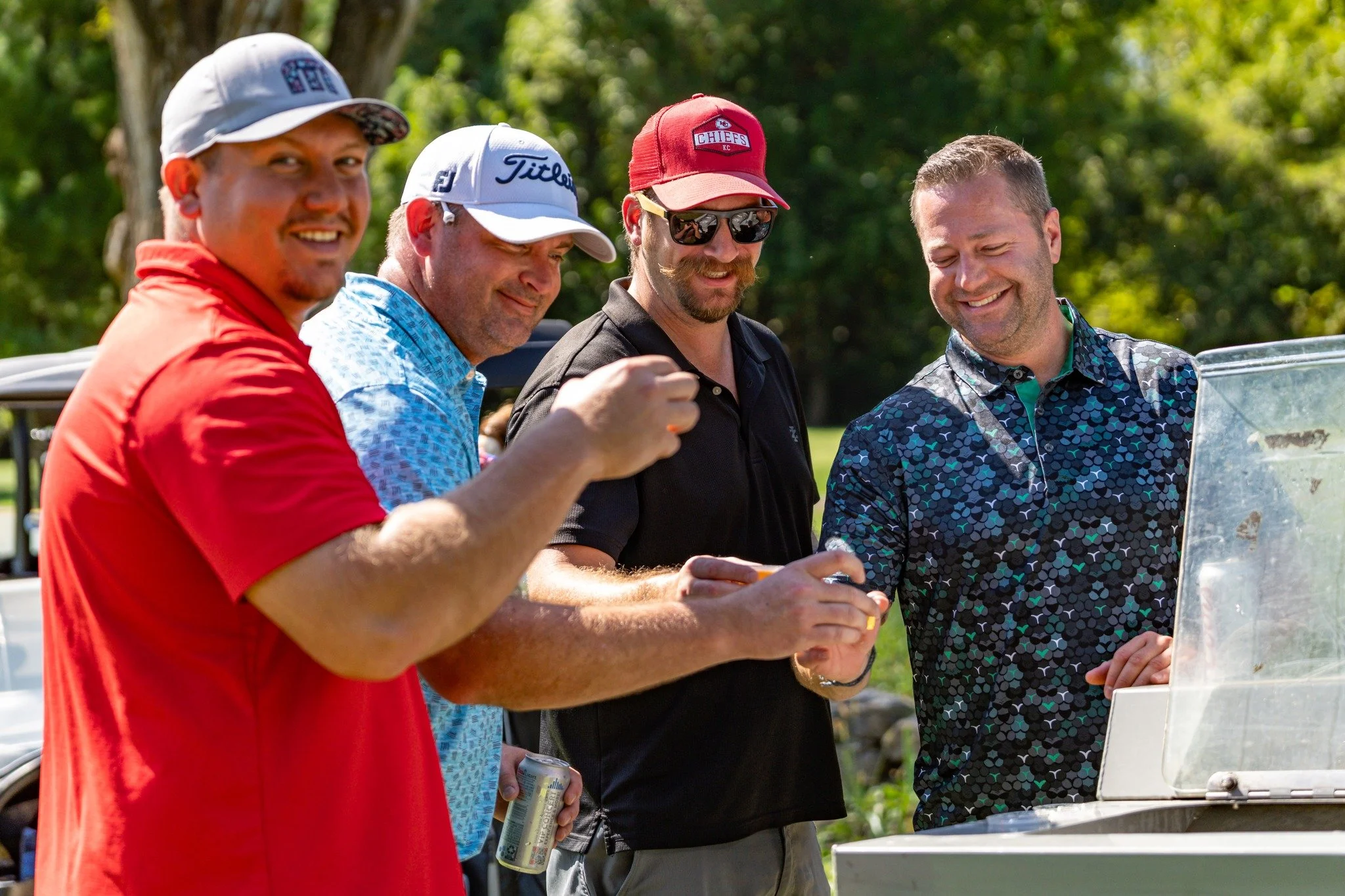 Four men smiling and enjoying a sunny day outside, standing near a small white outdoor heater or grill, with one holding a canned beverage and another wearing a red baseball cap and sunglasses.