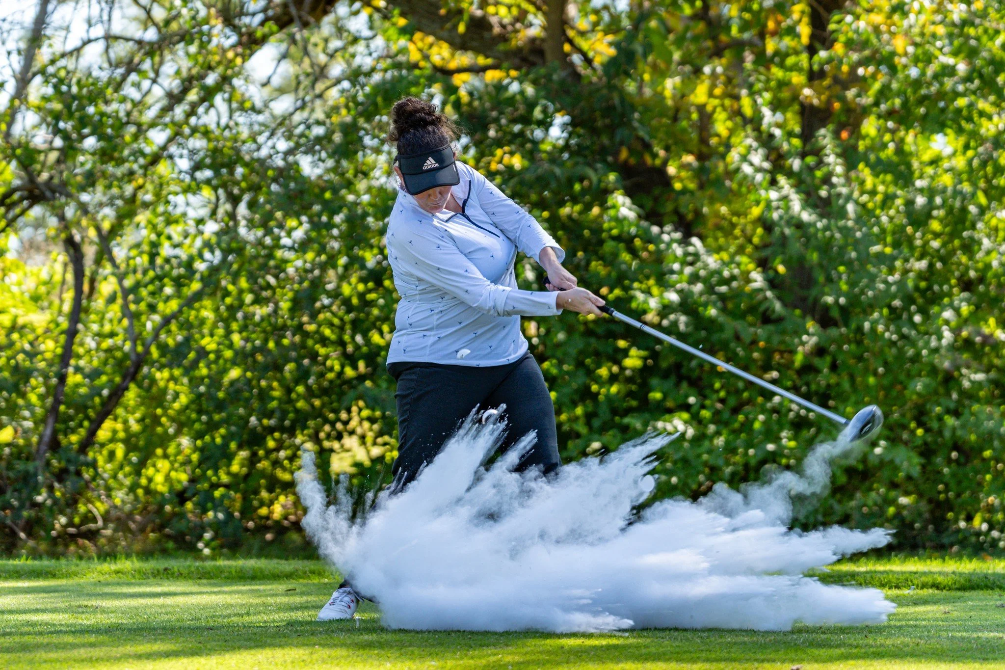 A woman in a white jacket and black pants hitting a golf ball, causing a large splash of water on a lush green golf course with trees in the background.
