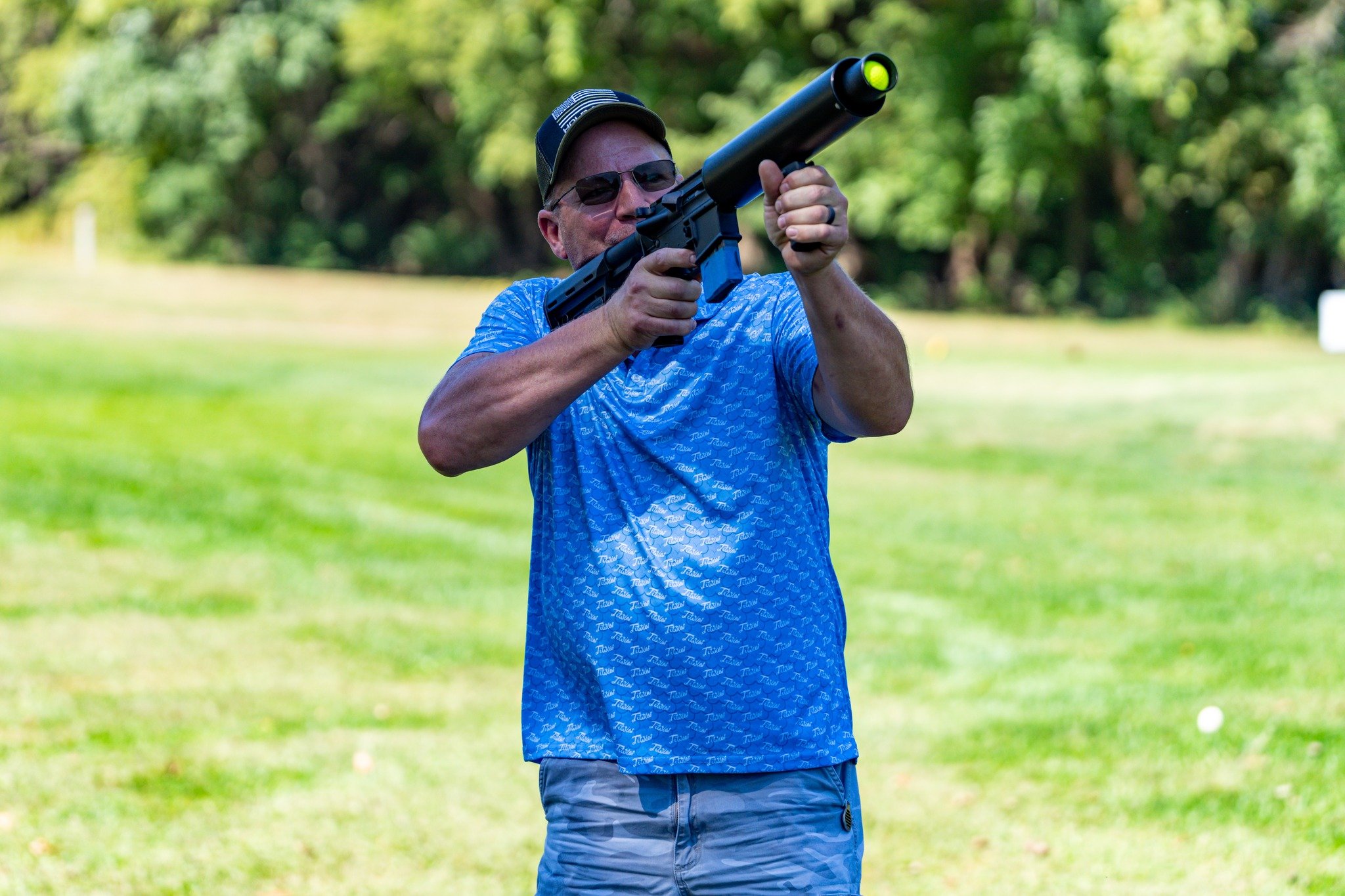 A man wearing sunglasses, a cap, and a blue shirt aiming a gun with a silencer on a grassy field.