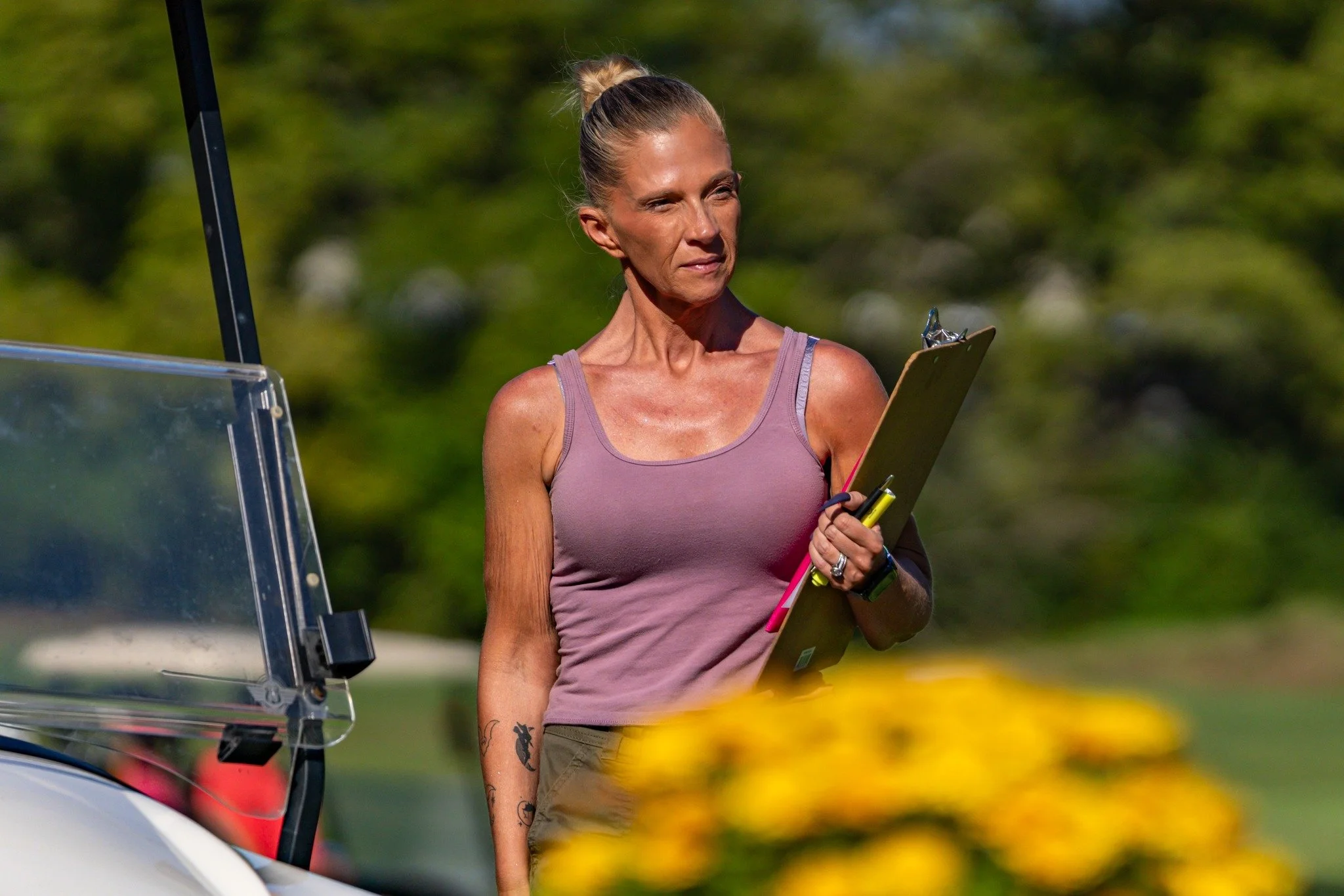 A woman with blonde hair in a bun, wearing a purple tank top, holding a clipboard and a pen, standing outdoors near what appears to be a golf cart. The background is blurred greenery and flowers.