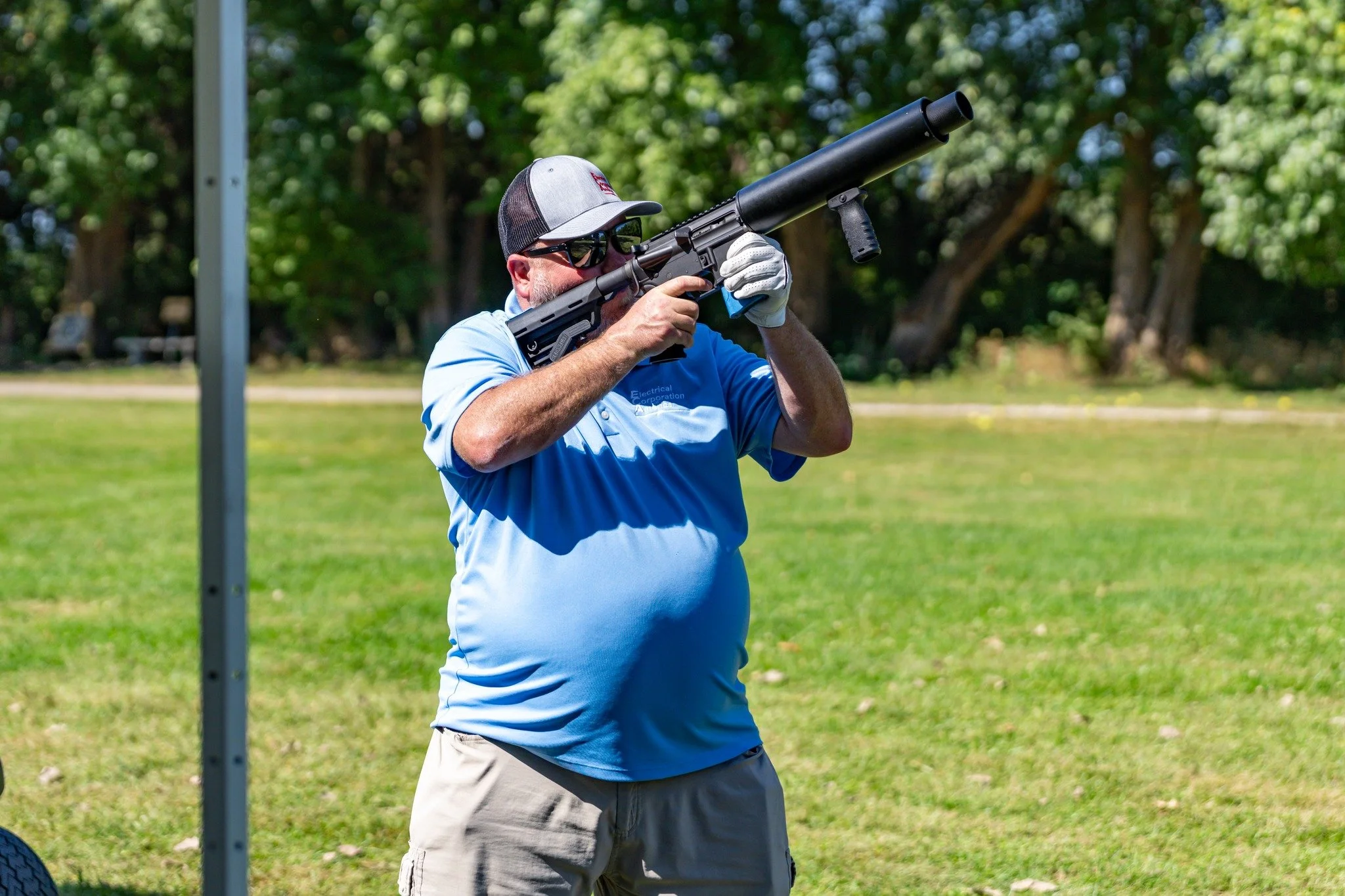 A man wearing a gray cap, sunglasses, and white gloves is aiming a black air rifle with a suppressor at a shooting range, standing on a grassy field with trees in the background.