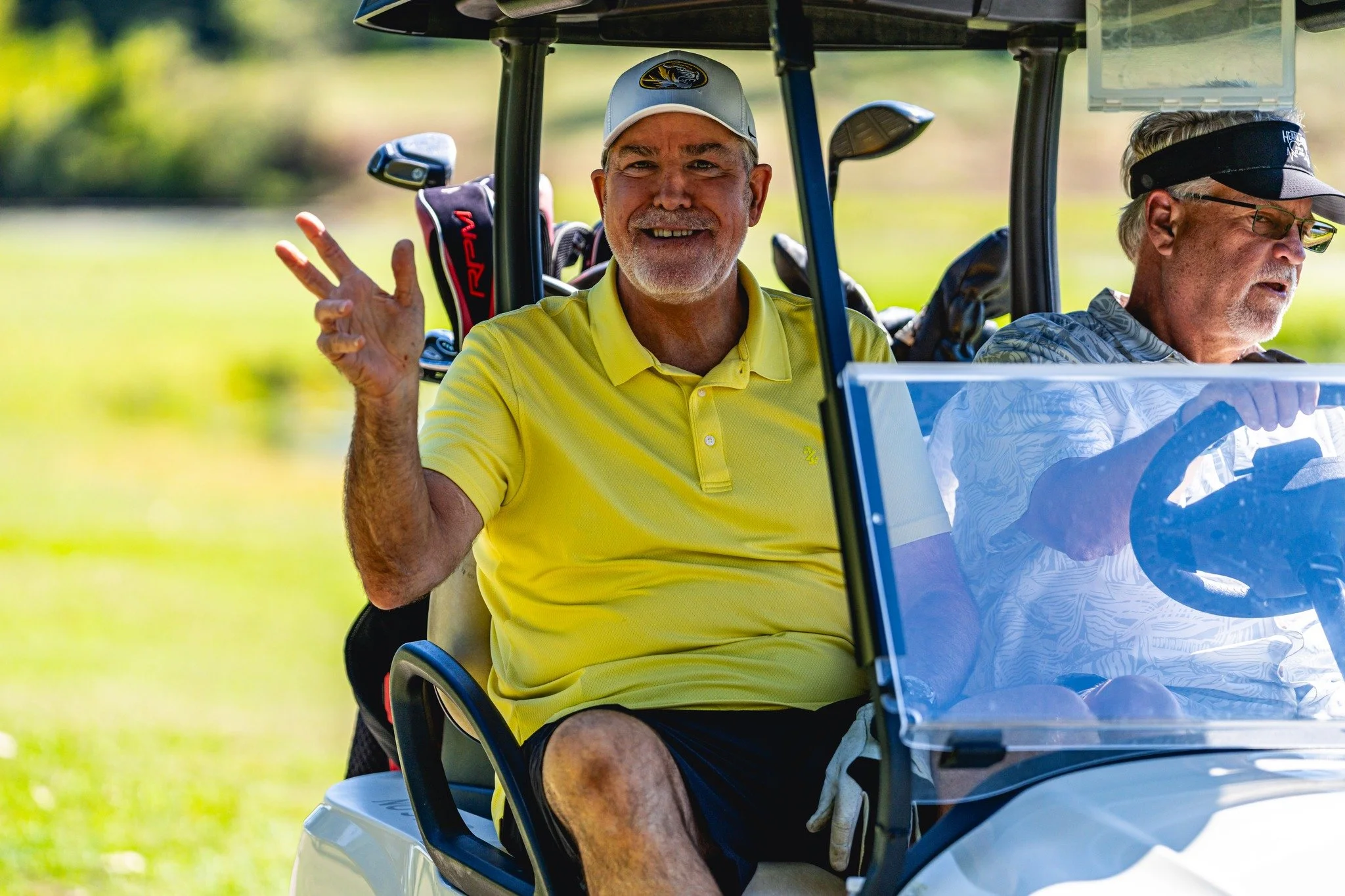 Two older men riding in a golf cart on a golf course. One man is wearing a yellow polo shirt and a golf cap, smiling and making a peace sign. The other man, wearing glasses and a visor, is driving the cart.
