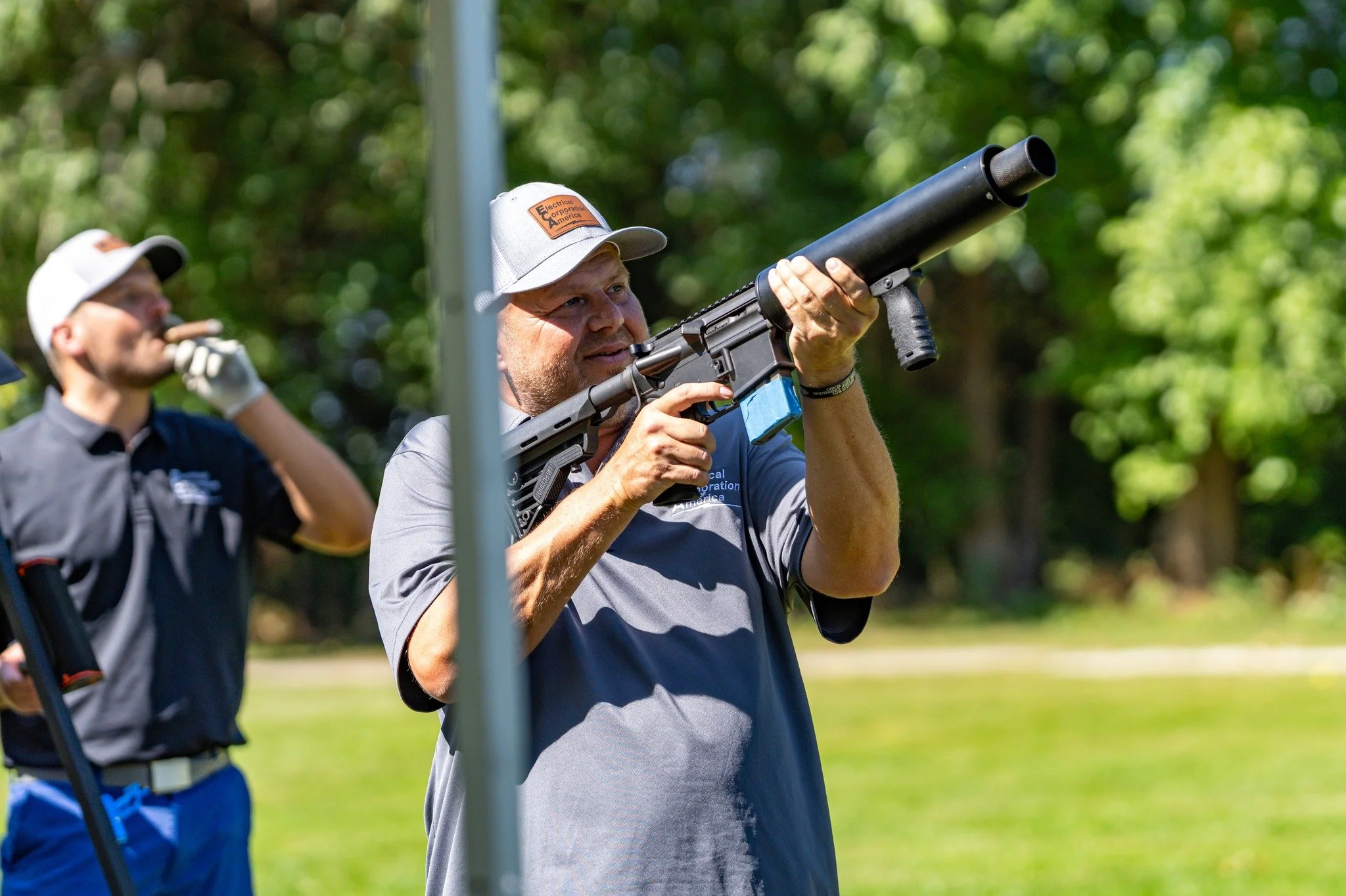 Man aiming a black gun with a silencer outdoors, with trees and grass in the background.