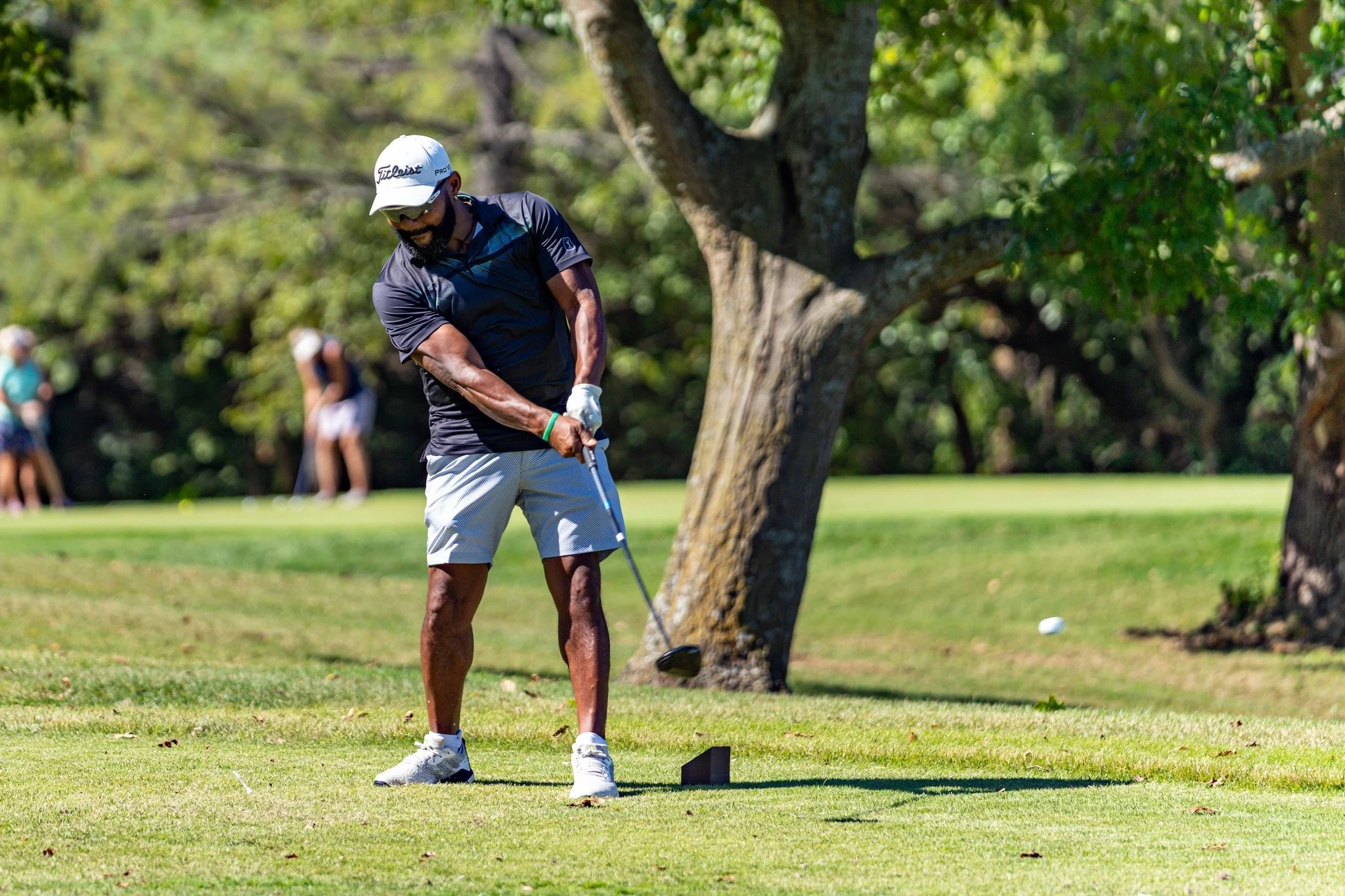A man playing golf on a lush green course, preparing to hit the golf ball near a tree, with a small group of people in the background.