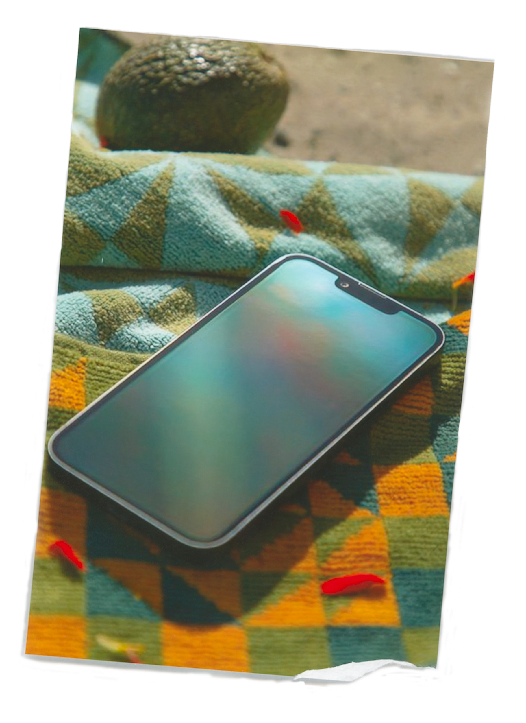 A smartphone with a reflective screen lies on a colorful, patterned textile. There is a round, textured object in the background, possibly a fruit or a decorative item.